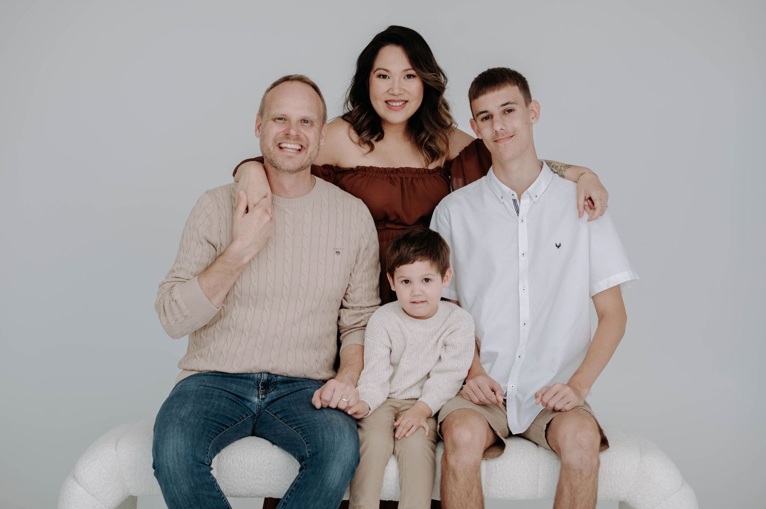 Studio family portrait with parents and children seated on white bench against gray background.