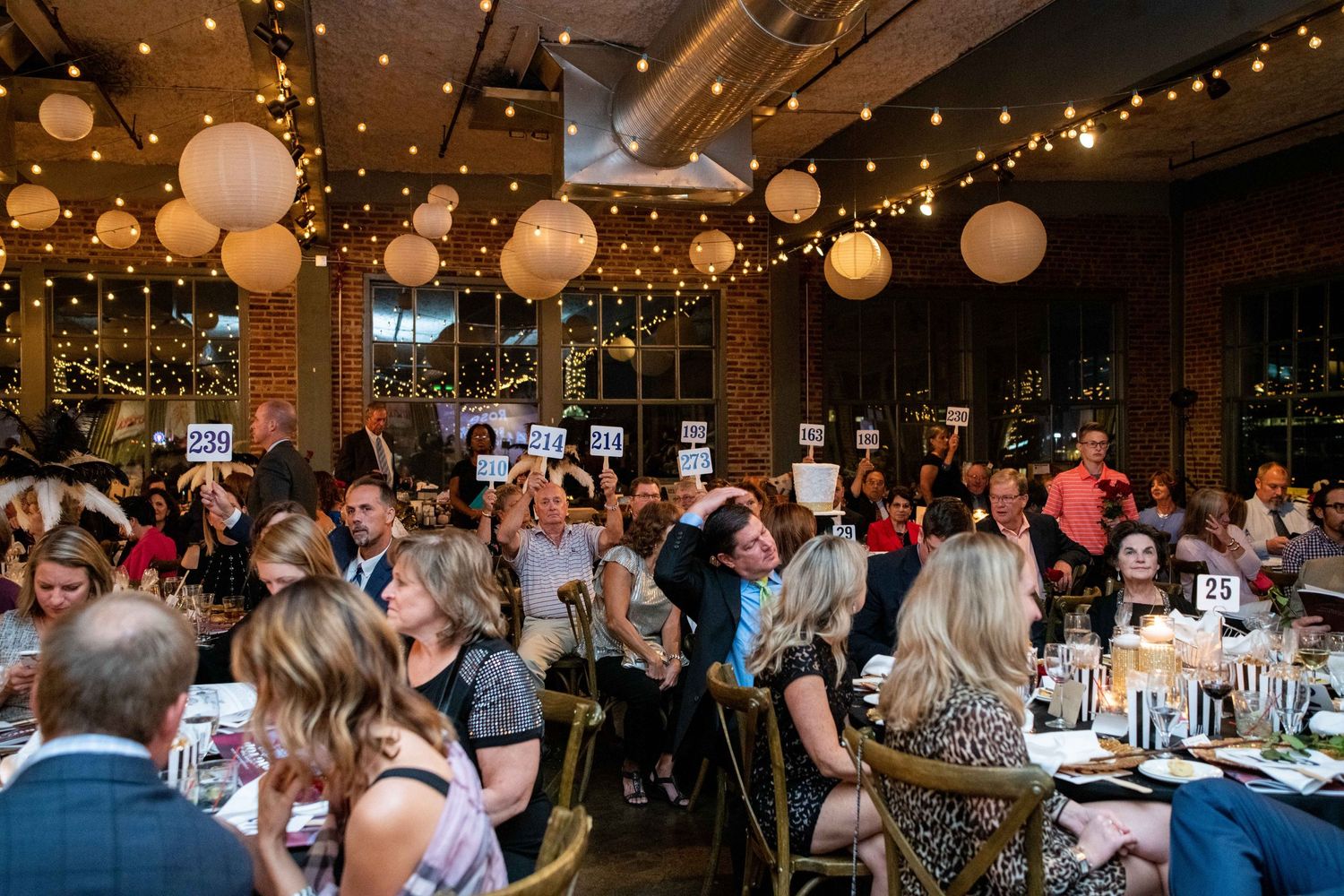 Festive indoor event venue with hanging paper lanterns and string lights as guests mingle at decorated dining tables.