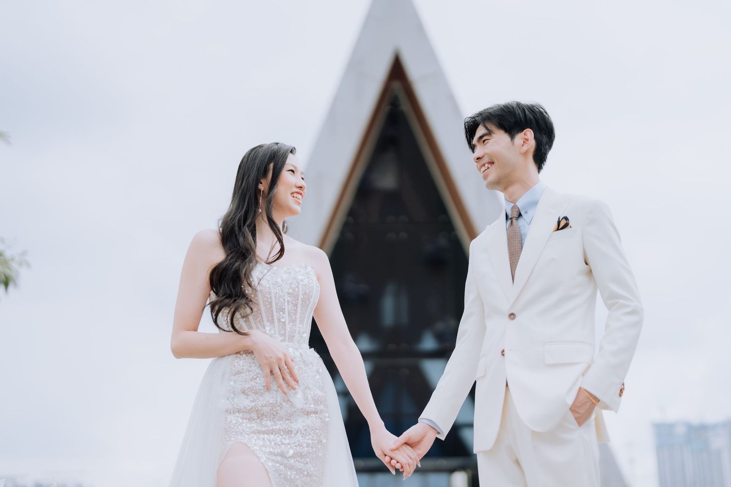 A couple in white wedding attire holding hands outside a modern chapel with an A-frame design.
