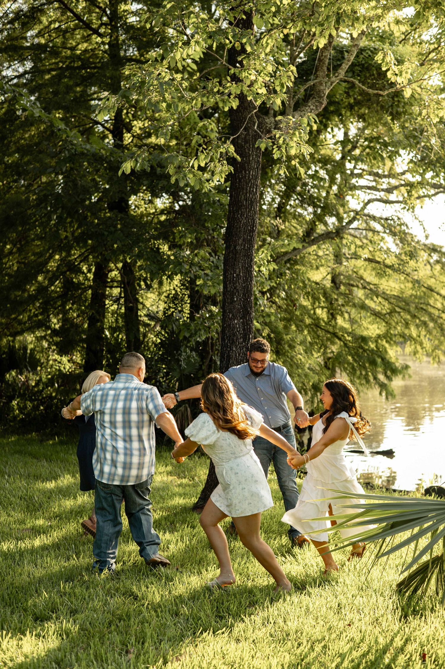 Family of five smiling together on green grass by a lakeside dock on a sunny day.