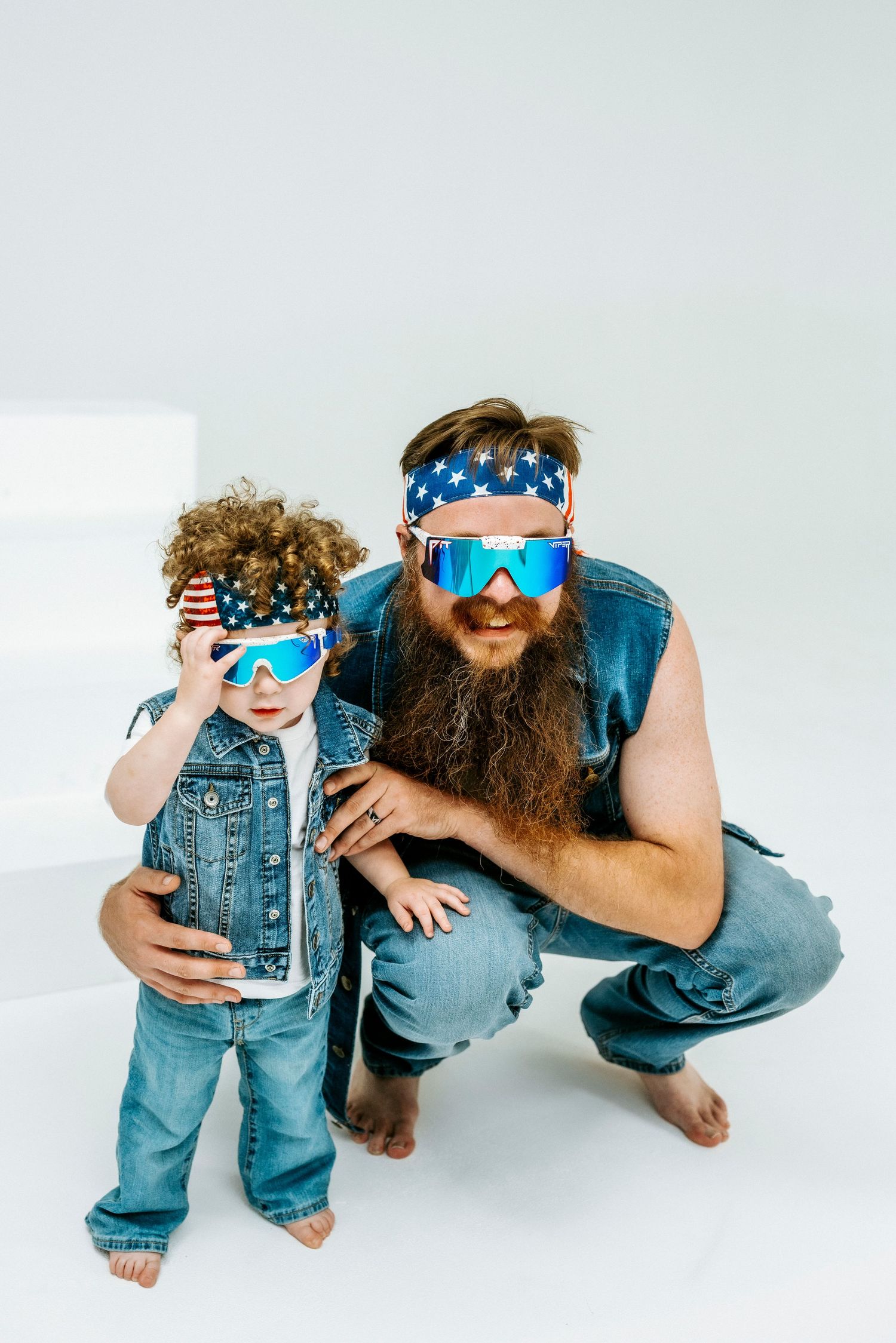 Bearded man and curly-haired child wearing matching American flag headbands and blue mirrored sunglasses in denim outfits.