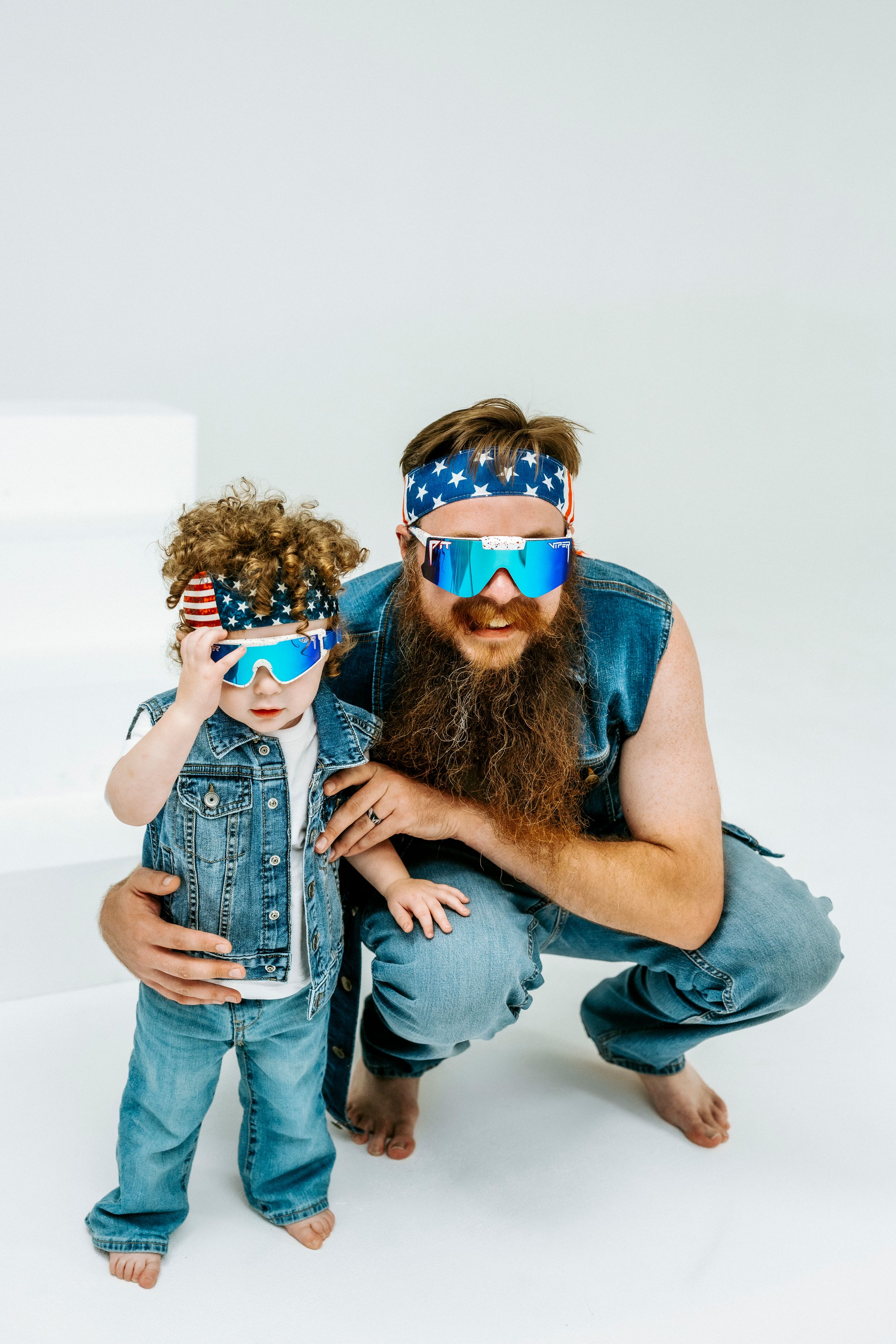 Bearded man and curly-haired child wearing matching American flag headbands and blue mirrored sunglasses in denim outfits.