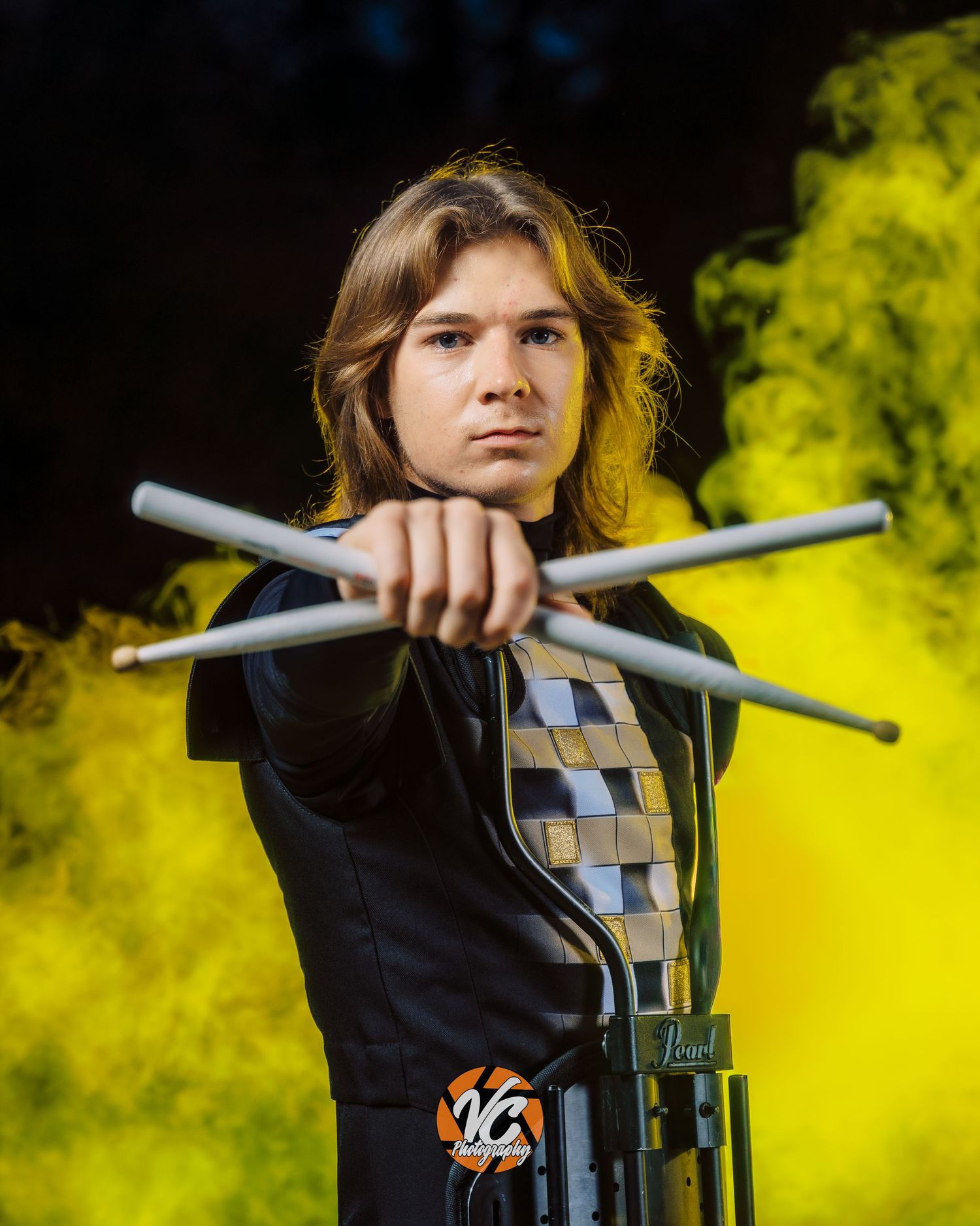 Young drummer poses with crossed drumsticks against vibrant yellow smoke background in a dramatic senior portrait photo.