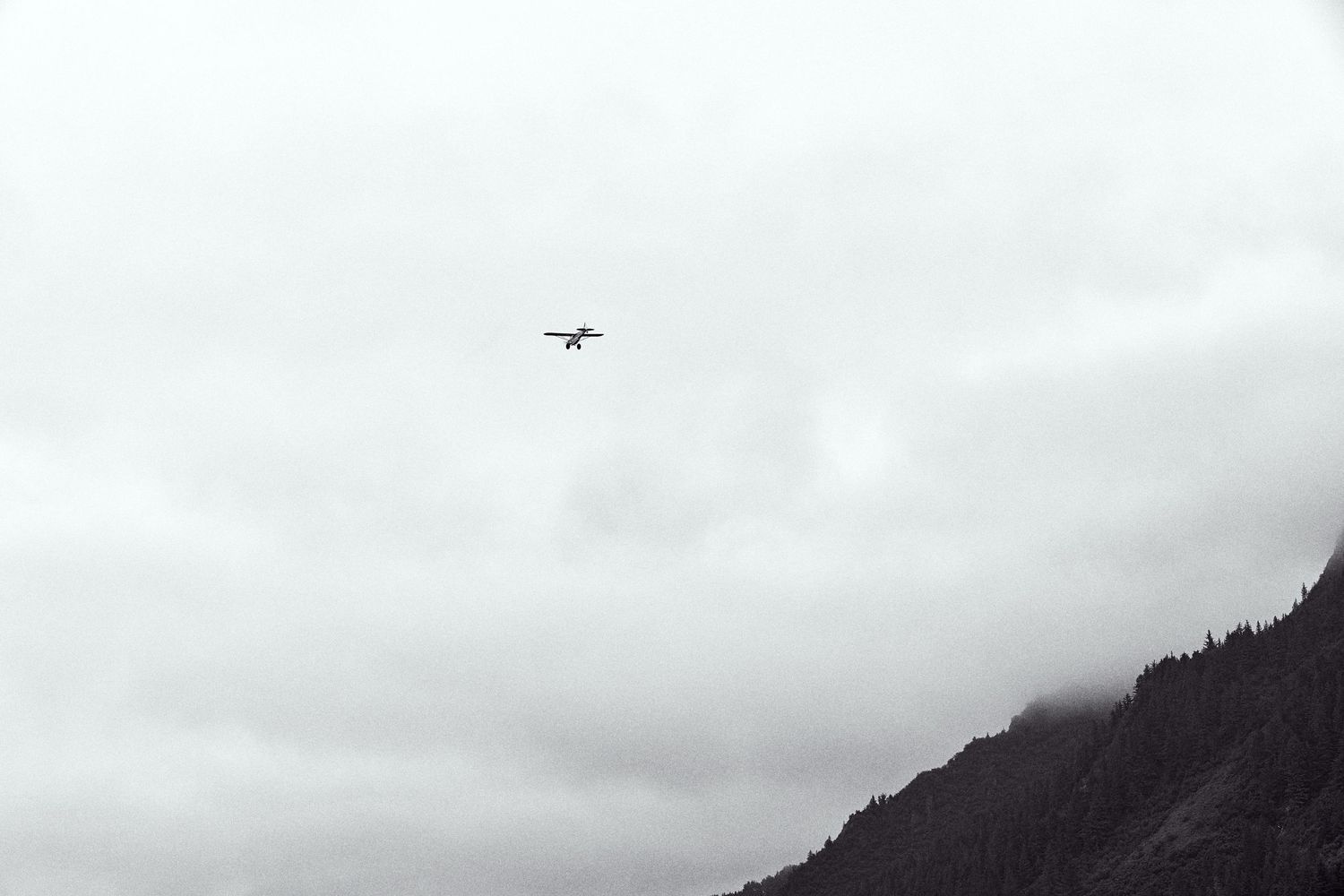 A small airplane flies through overcast cloudy skies above misty mountain peaks in a moody black and white landscape photo.