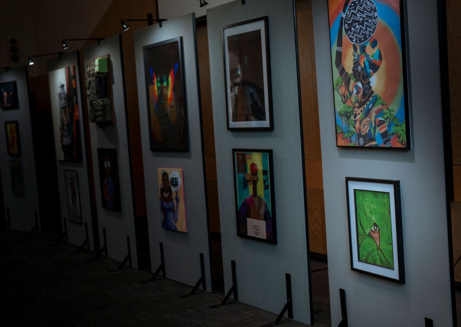 Dimly lit art gallery hallway displaying colorful framed paintings and photographs on white display panels.
