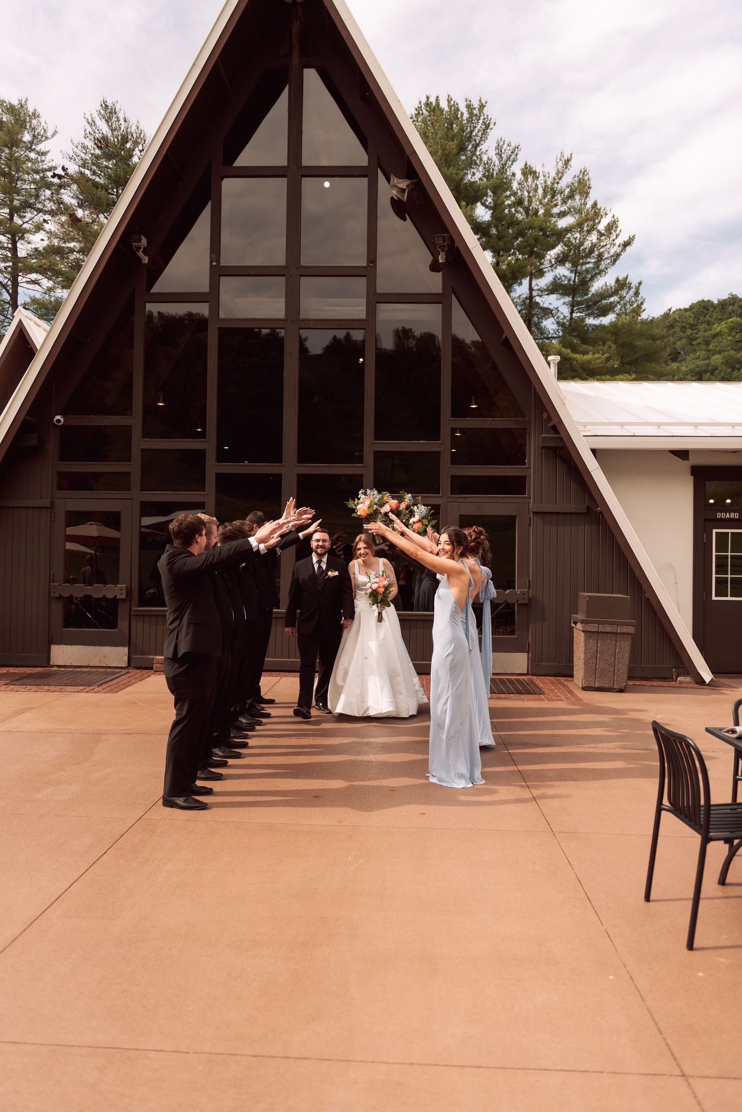 Bride and groom exit A-frame chapel while wedding party forms celebratory arch outdoors.