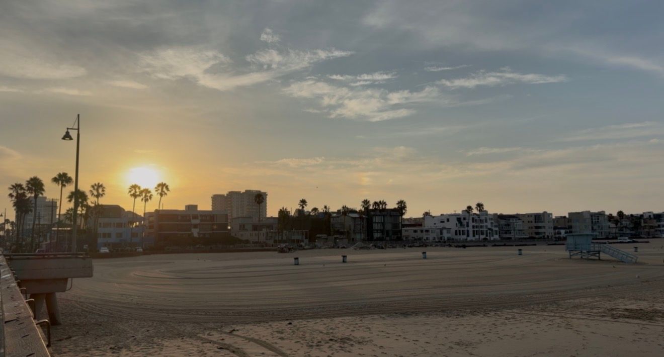 Sunset over Venice Beach boardwalk with palm trees, sandy shore, and coastal buildings glowing in golden evening light.