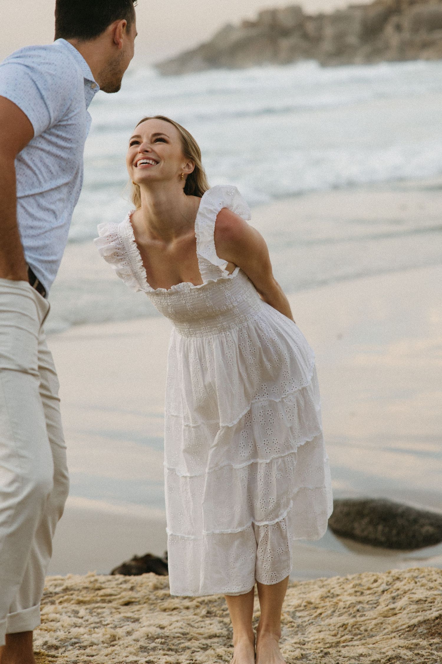 A smiling woman in a white ruffled dress stands barefoot on a sandy beach with a man in a gray shirt.