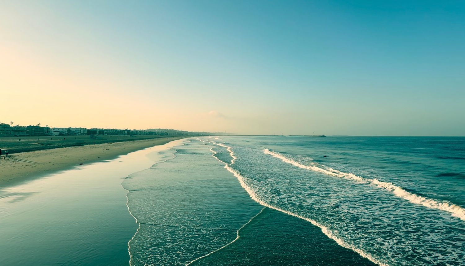 Aerial view of a serene beach at sunset with gentle waves, golden sand, and a clear blue sky over the ocean.