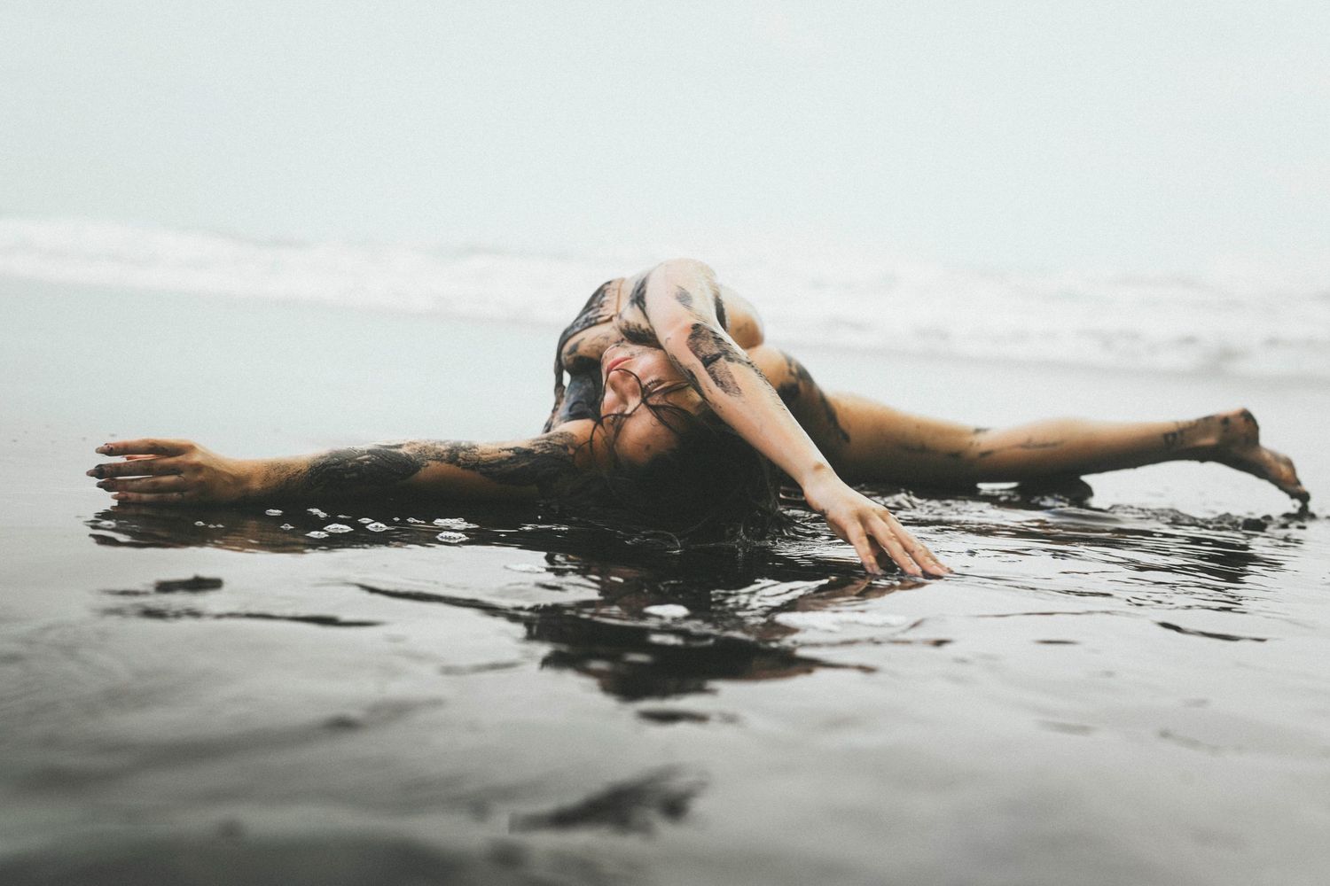 Woman in bikini lying on dark sandy beach with ocean waves crashing around her under dramatic cloudy skies.