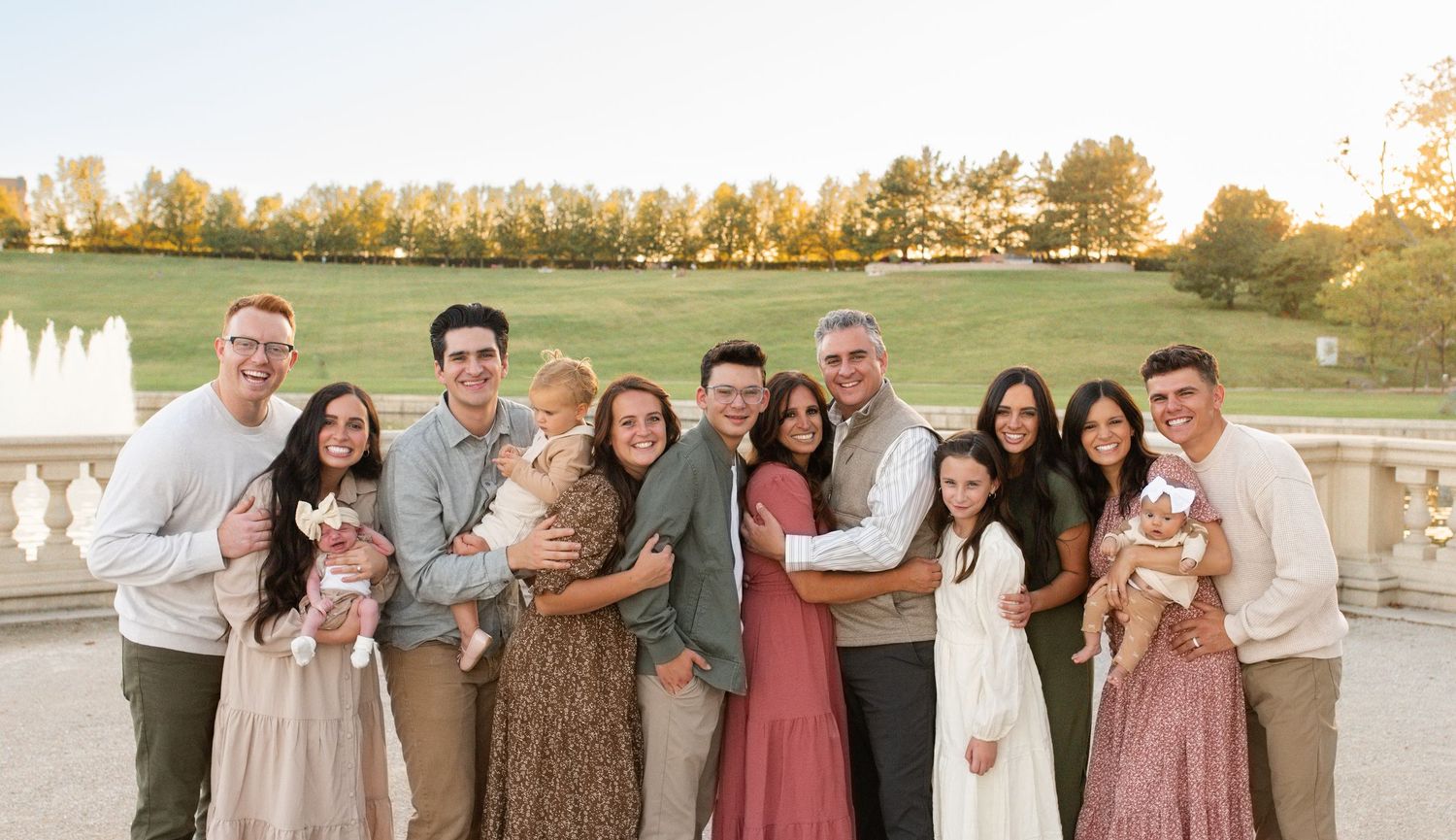 Large family group portrait outdoors near fountain, wearing coordinated earth tones, smiling together at golden hour.