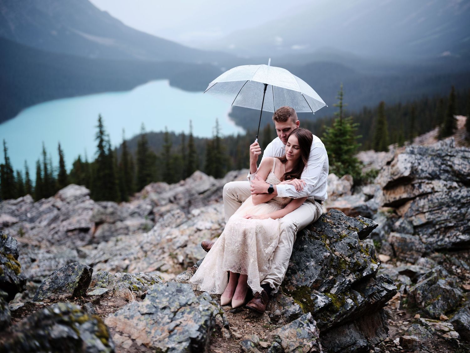 Couple sharing an umbrella on rocky mountain overlook above turquoise lake during rainy elopement session.