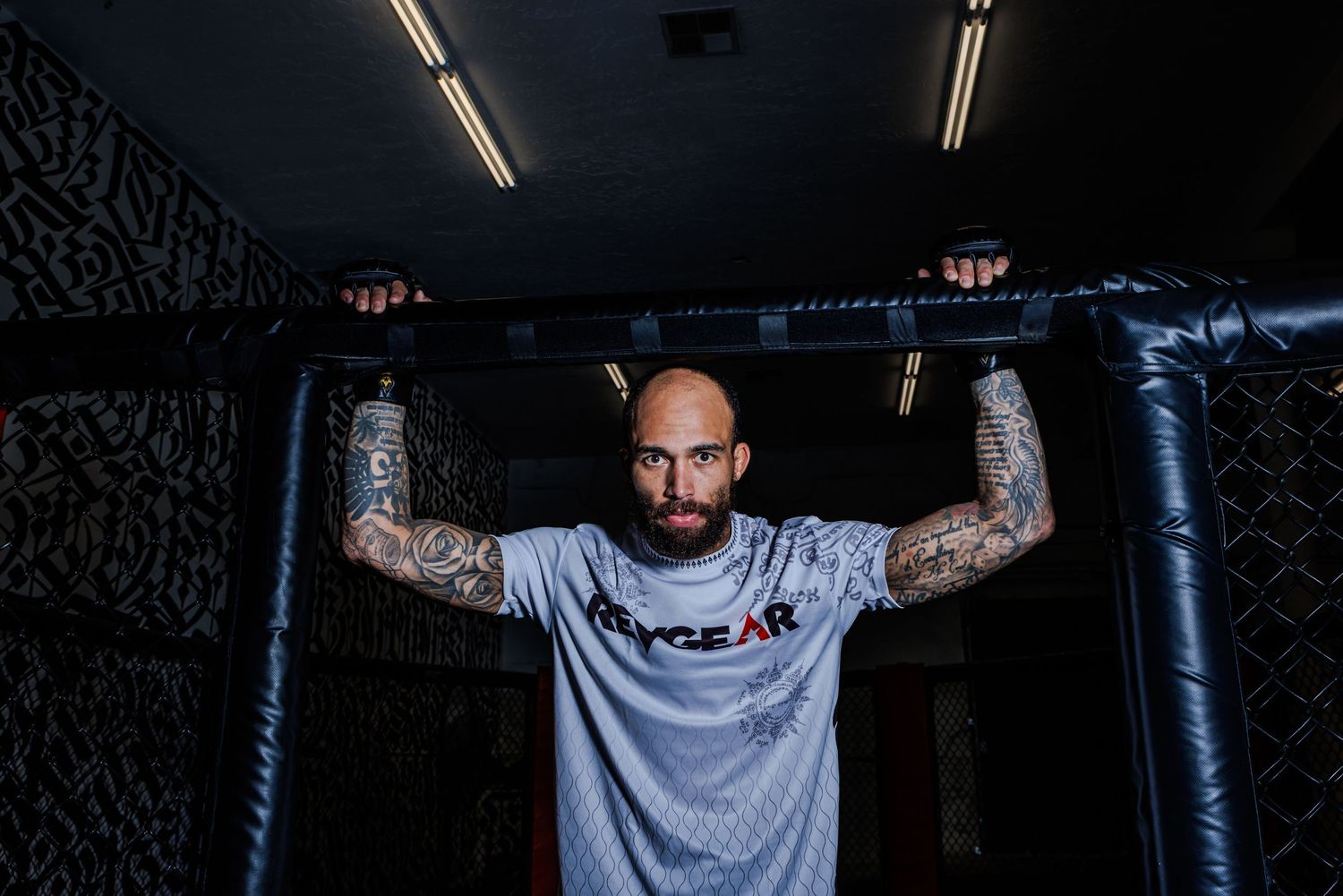 Athletic person with tattoos grips a pull-up bar in a dimly lit gym while wearing a light gray t-shirt.