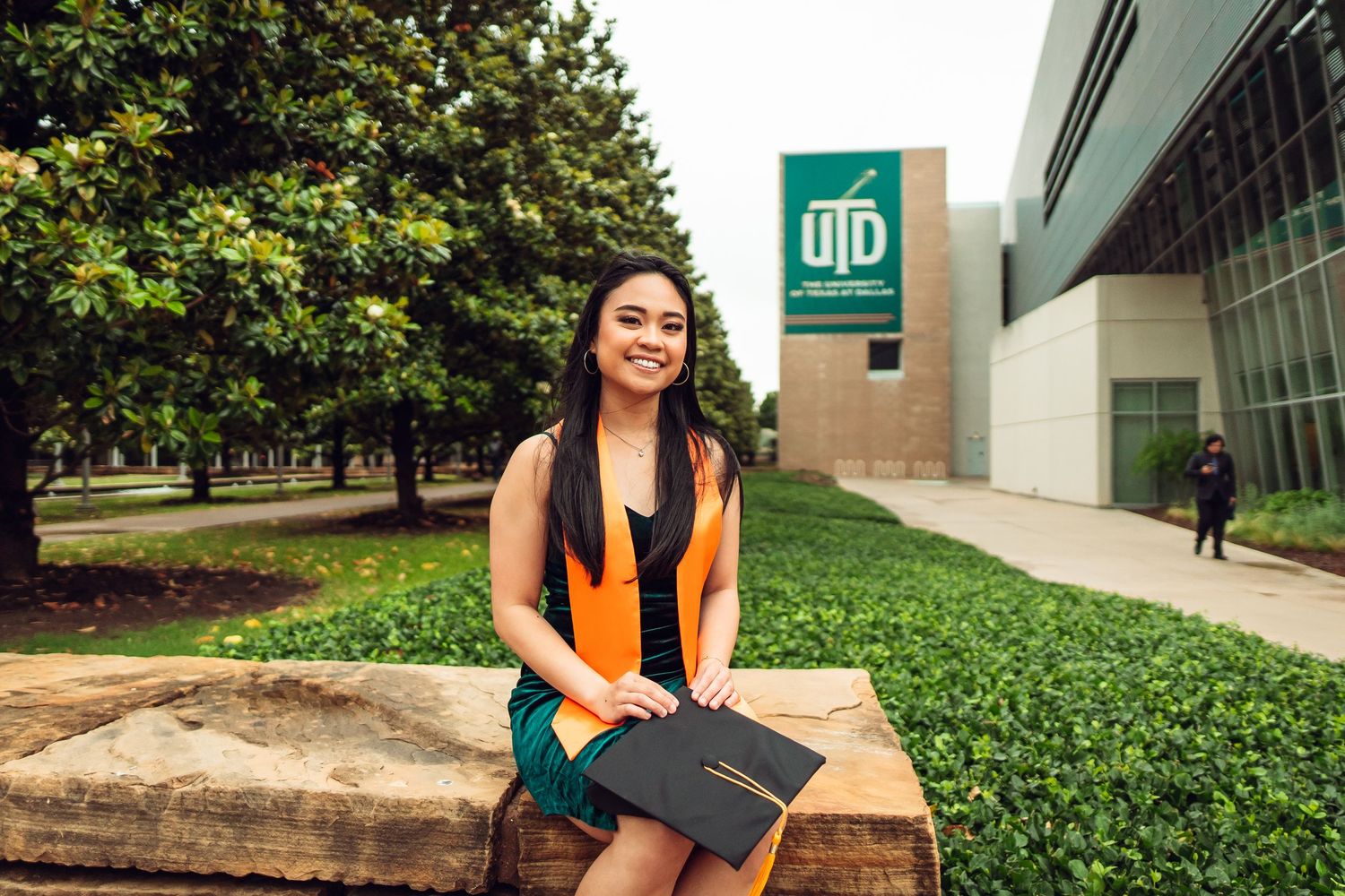 Graduate posing in orange stole and black gown on campus with green landscaping.
