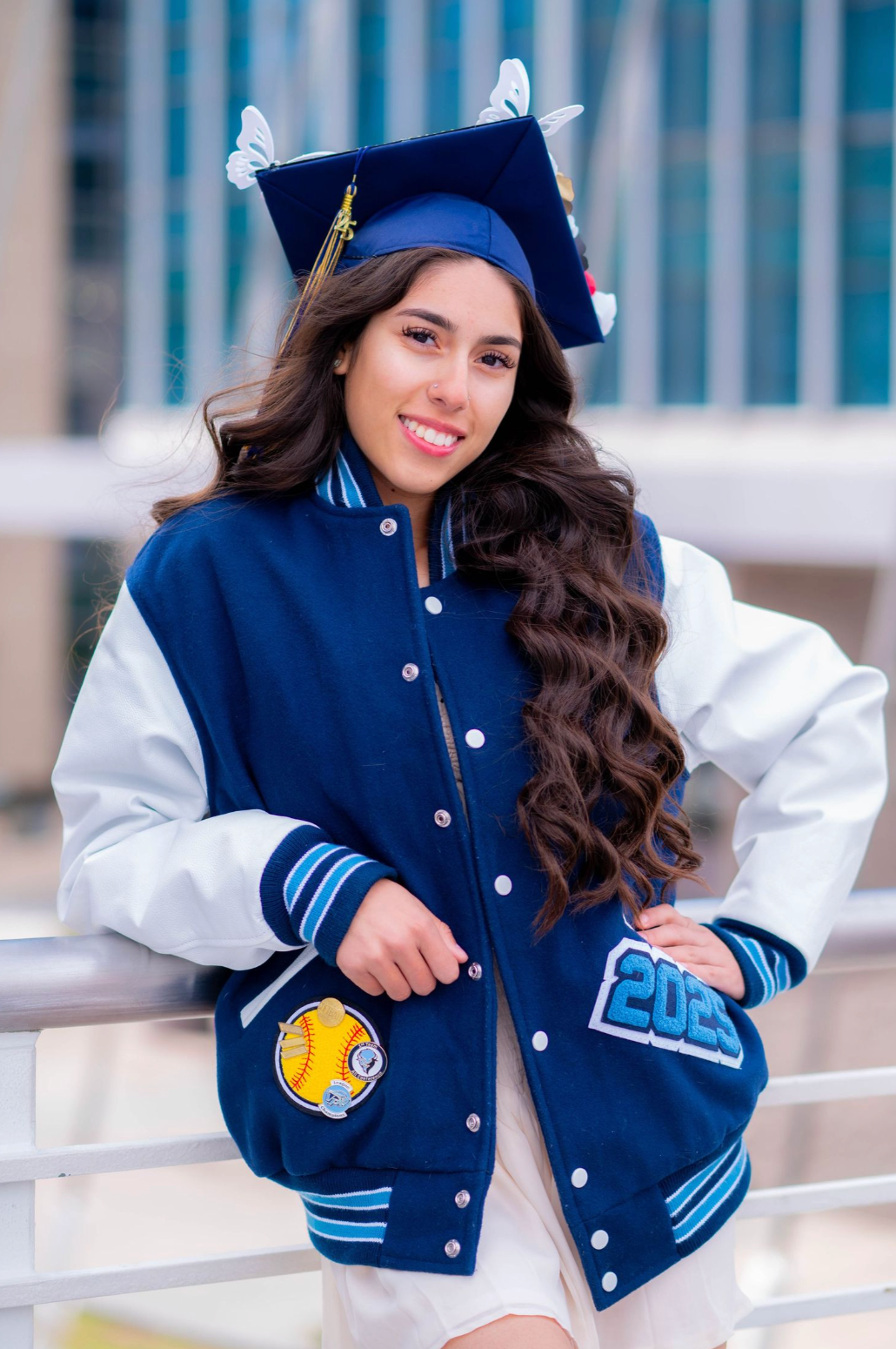 A graduate wearing a blue and white varsity jacket and cap poses on outdoor steps.