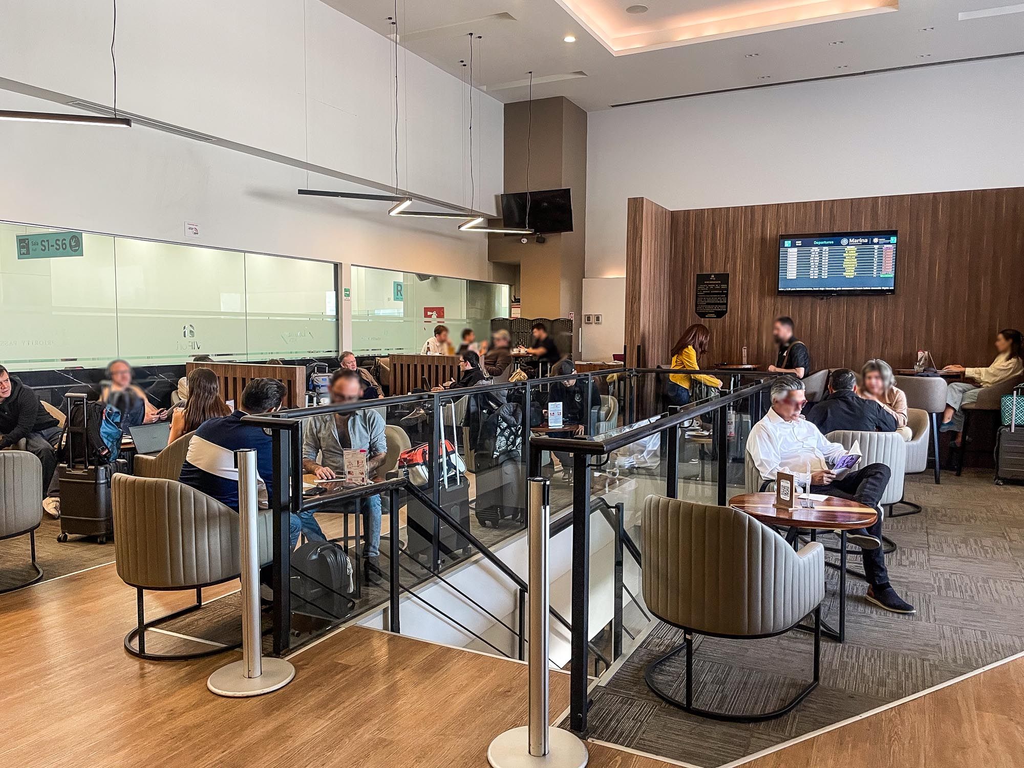 Modern airport lounge interior with glass partitions, wooden walls, and travelers seated at various workstations.