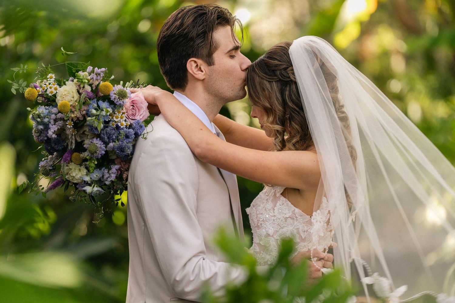 A bride and groom share a romantic kiss outdoors, surrounded by lush greenery, as she holds a colorful wedding bouquet.