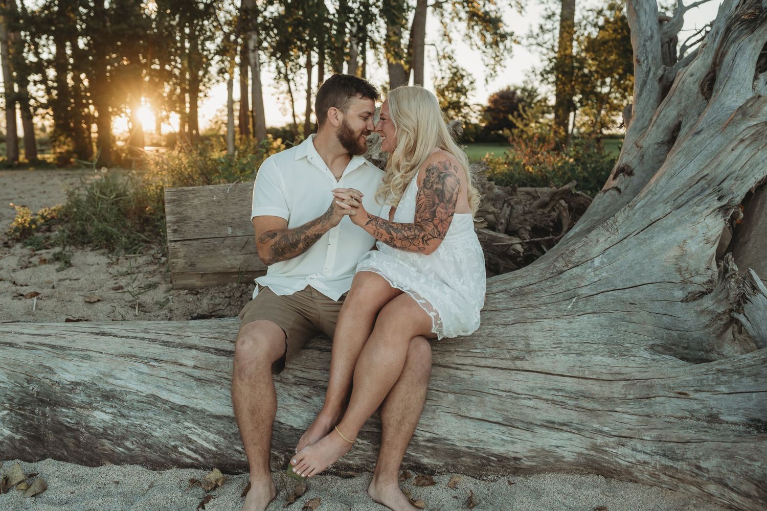 Couple sitting on driftwood at sunset on a sandy beach, sharing a romantic moment surrounded by trees.