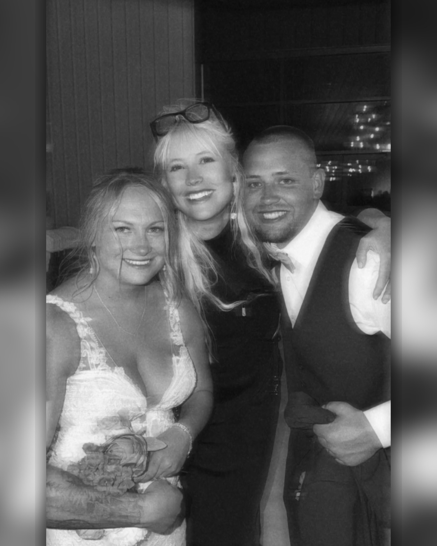 Black and white photo of three smiling people posing together at a formal event, two blonde women and a man in a vest.