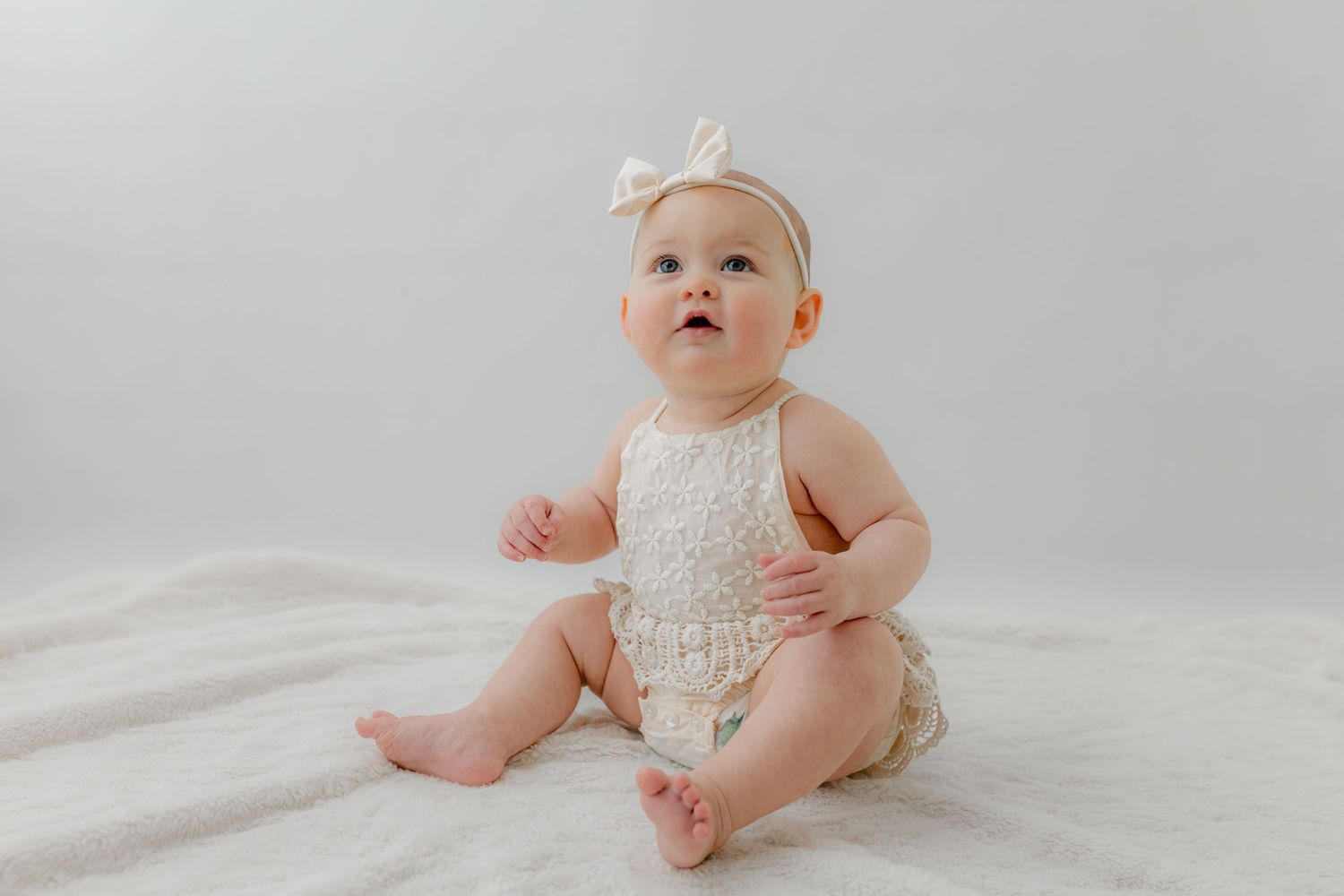A cute baby girl in a cream lace romper and white bow headband sits on a white blanket looking up.