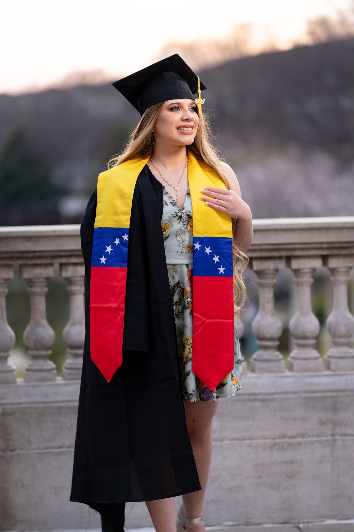 Young woman in graduation cap and gown wearing Venezuelan flag stole, smiling outdoors near stone balustrade.