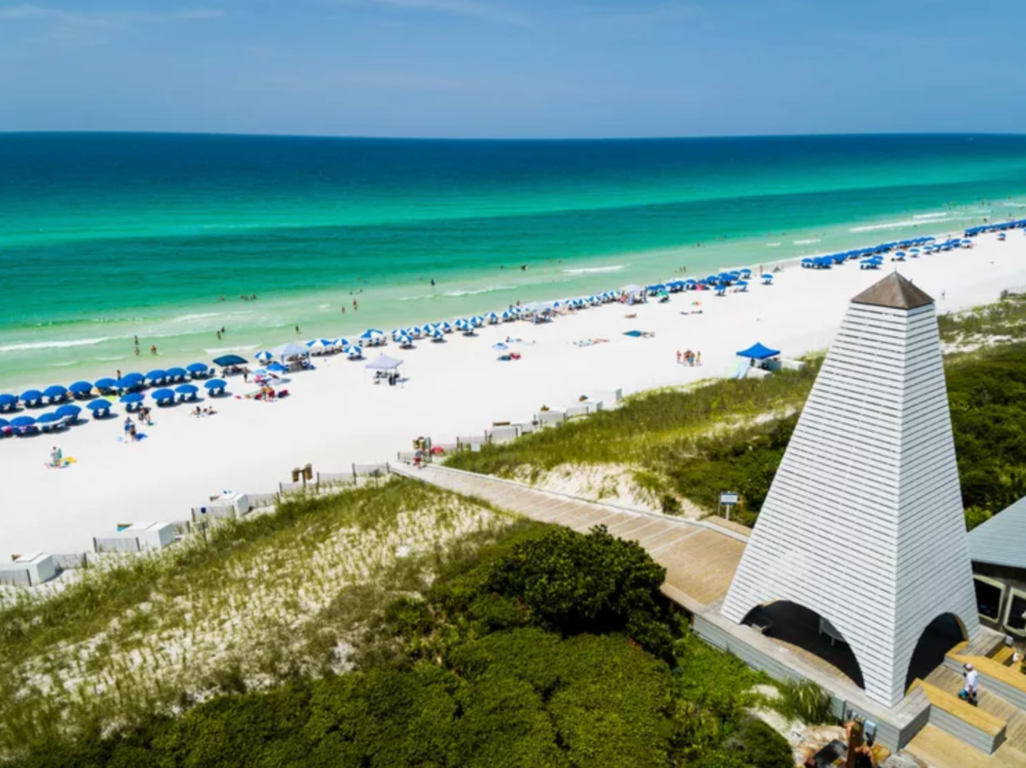 Aerial view of a white sand beach with turquoise water, blue umbrellas, and a pyramid-shaped structure in Destin, Florida.
