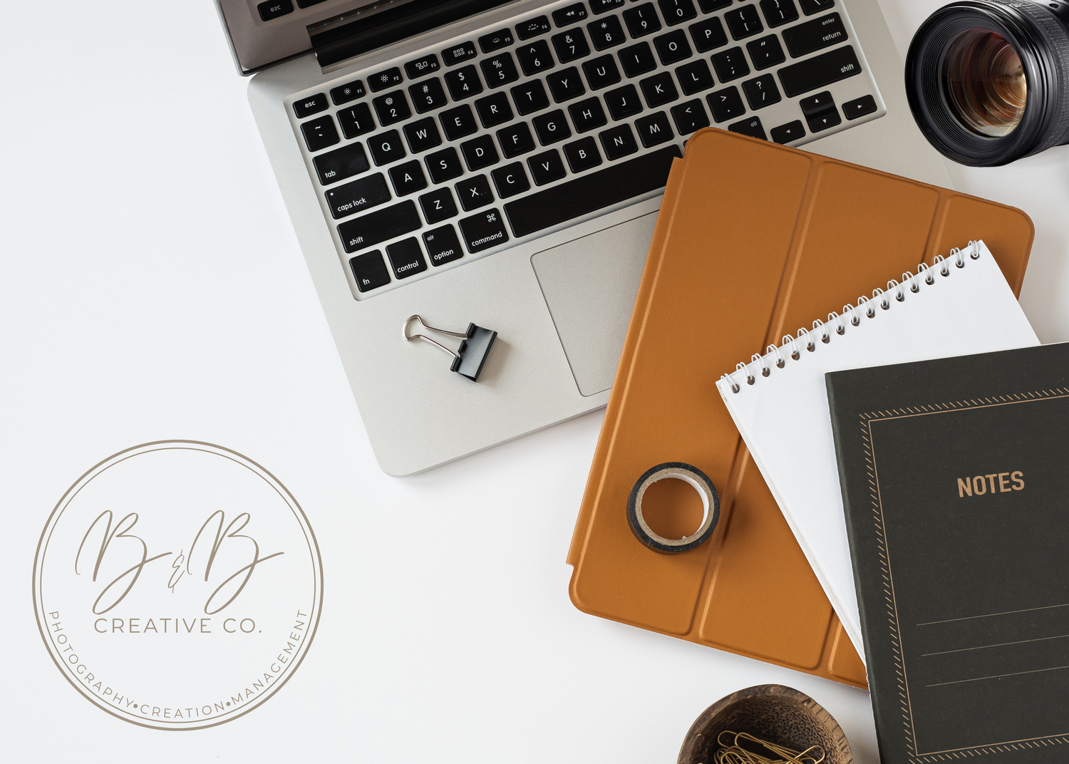 Flatlay of a laptop, tan iPad case, spiral notepad, black notes journal, camera lens, and binder clip on white background.
