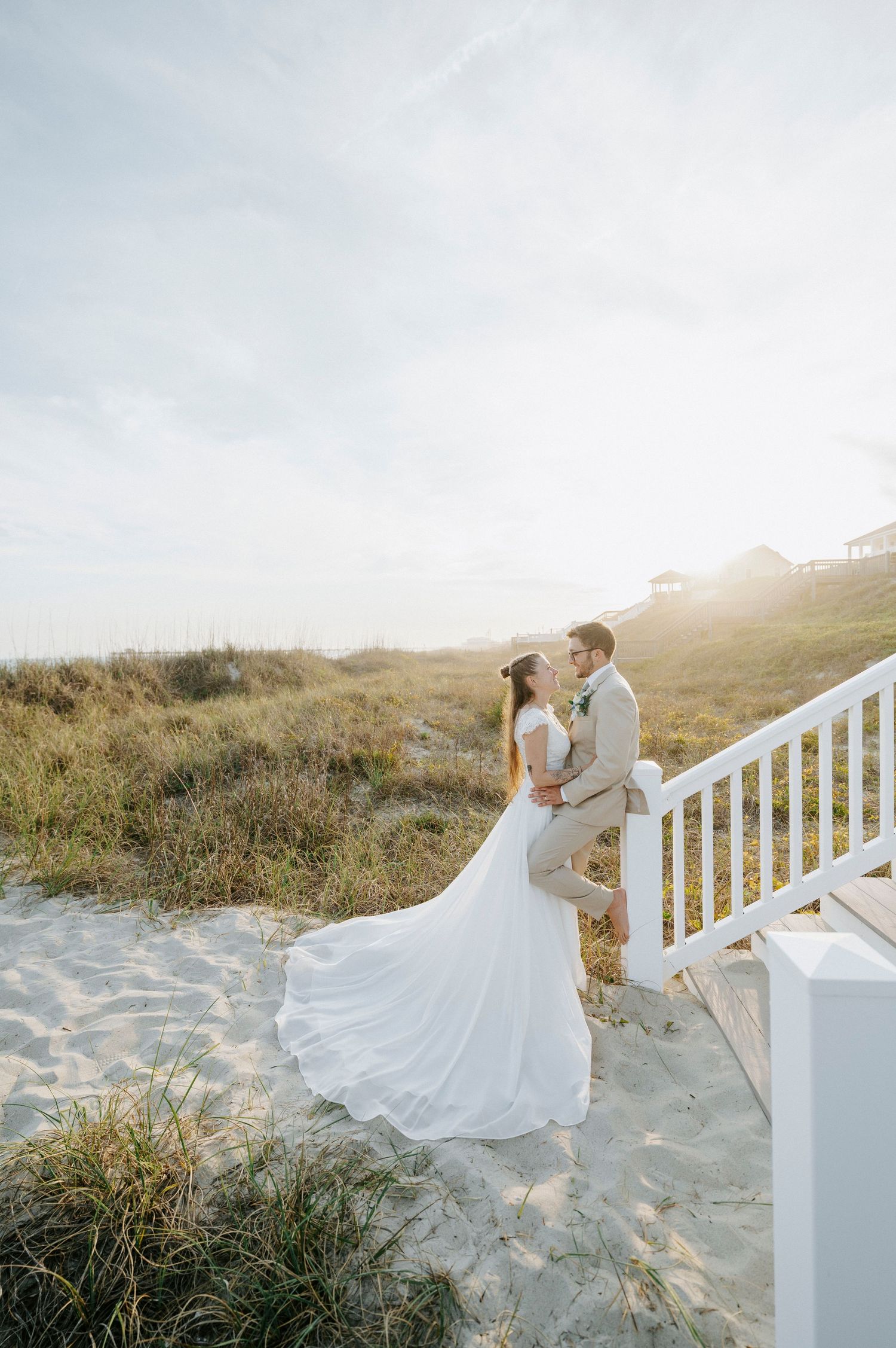 Bride and groom embrace on sandy beach stairs at sunset, her white gown trailing on the sand.
