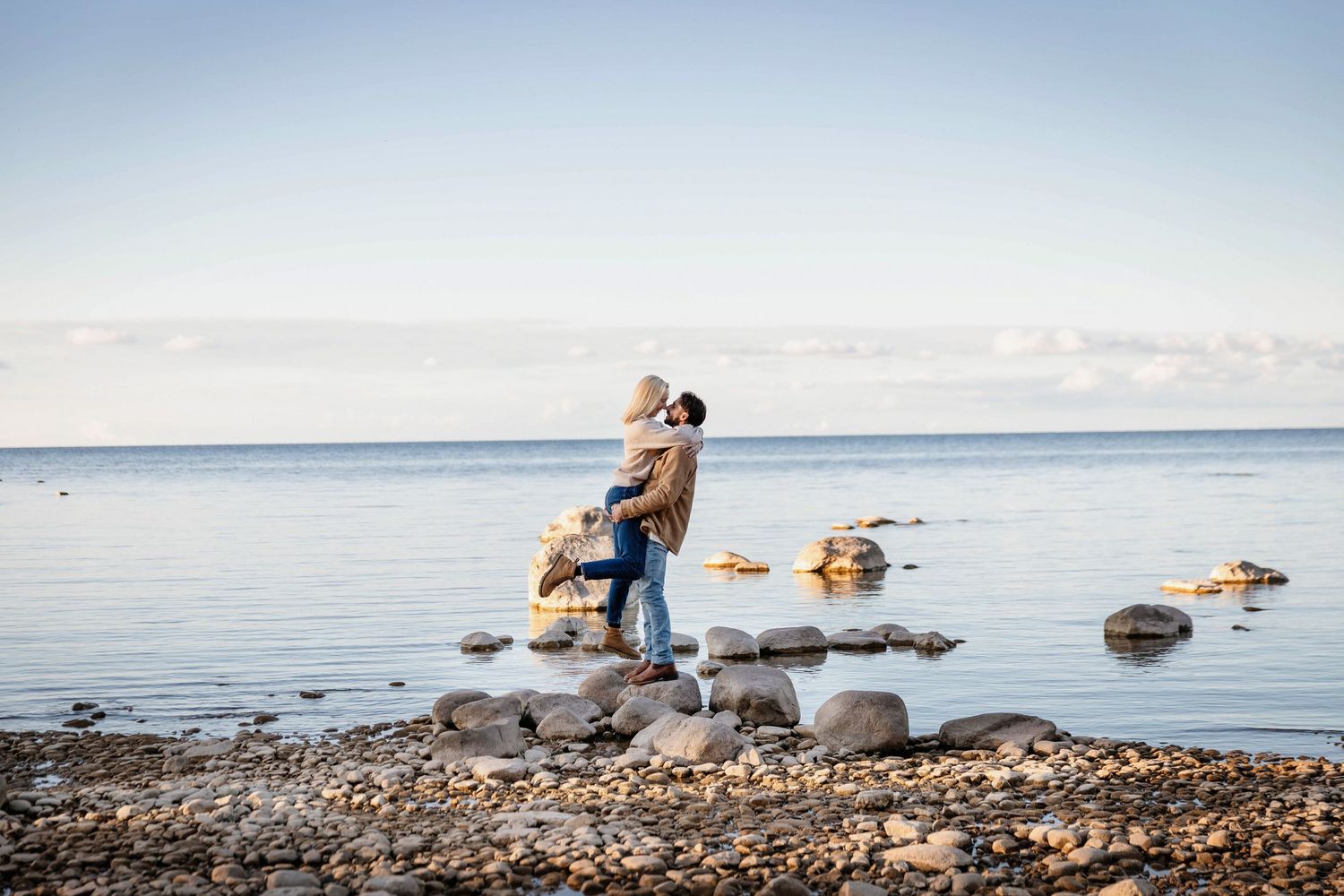 Couple embracing on rocky shoreline with calm ocean waters and clear blue sky in background.