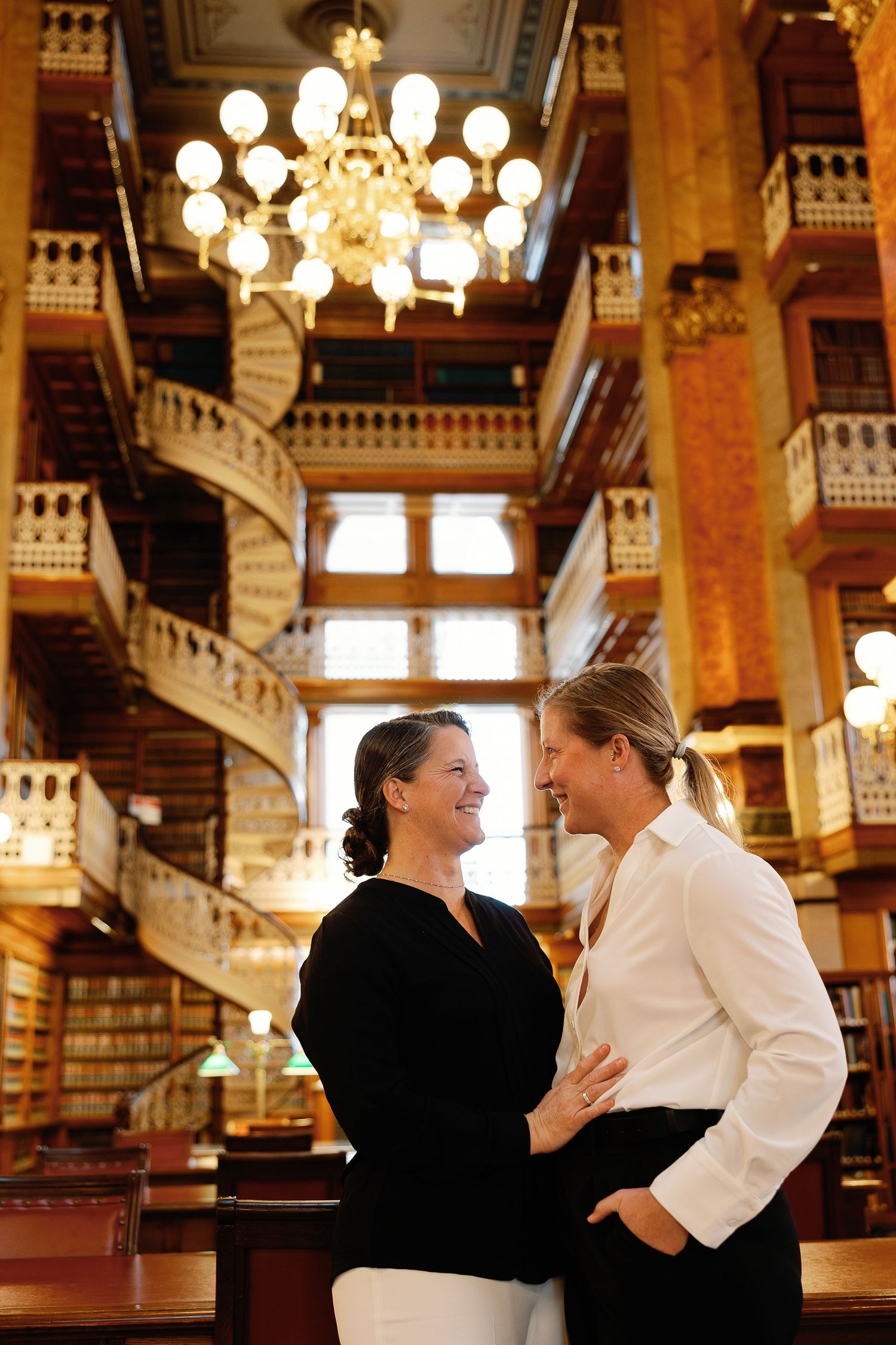 Two women in white and black dress shirts face one another, smiling and embracing in, an ornate library at the Iowa State Capitol.