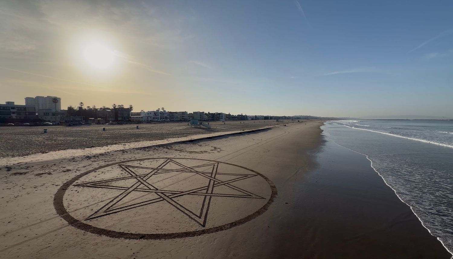 Large pentagram drawn in sand on a sunny beach with ocean waves and city skyline in the background.