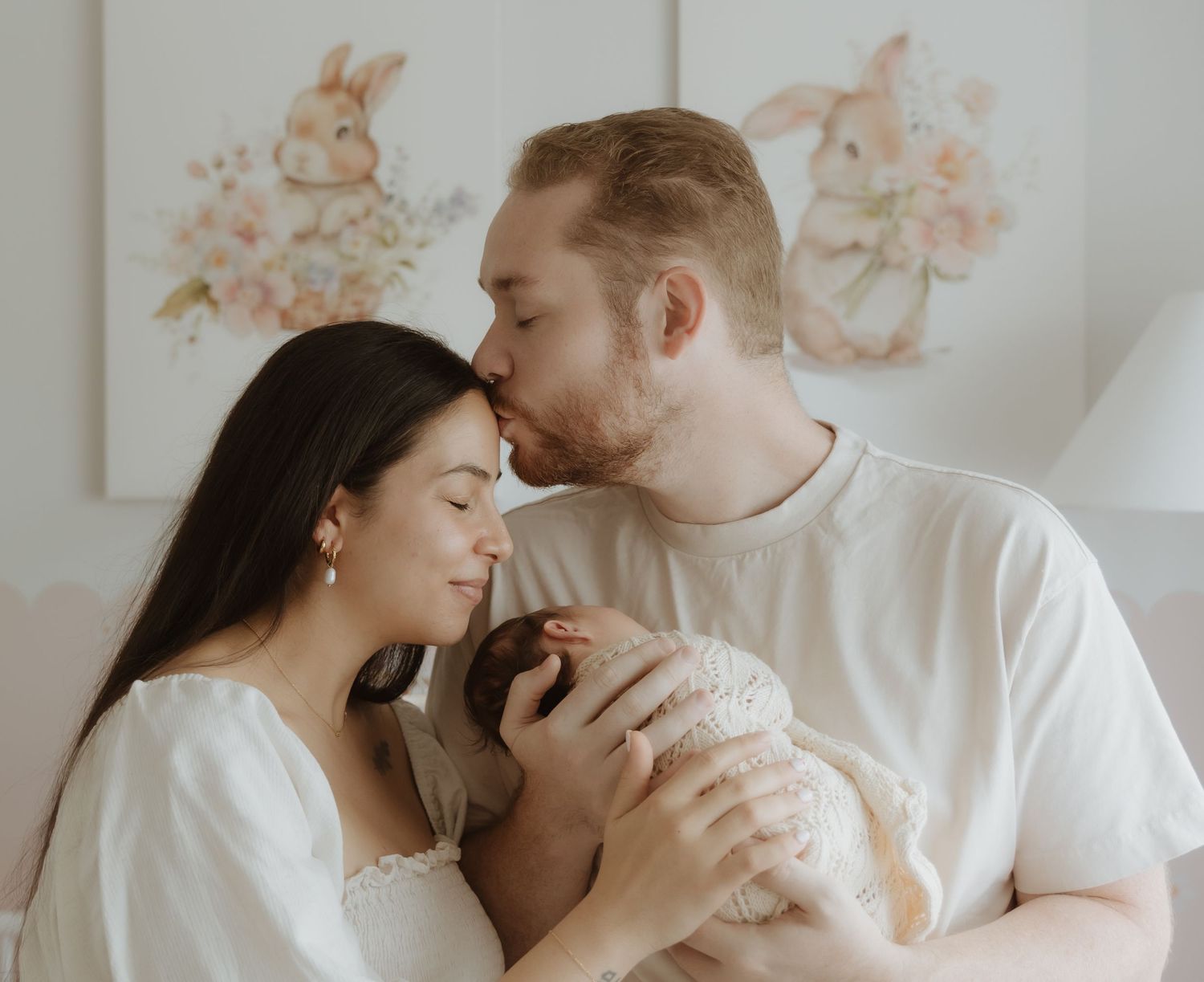 A couple tenderly kisses their newborn baby in a soft, pastel nursery with bunny wall art. Based in Lake Macquarie NSW