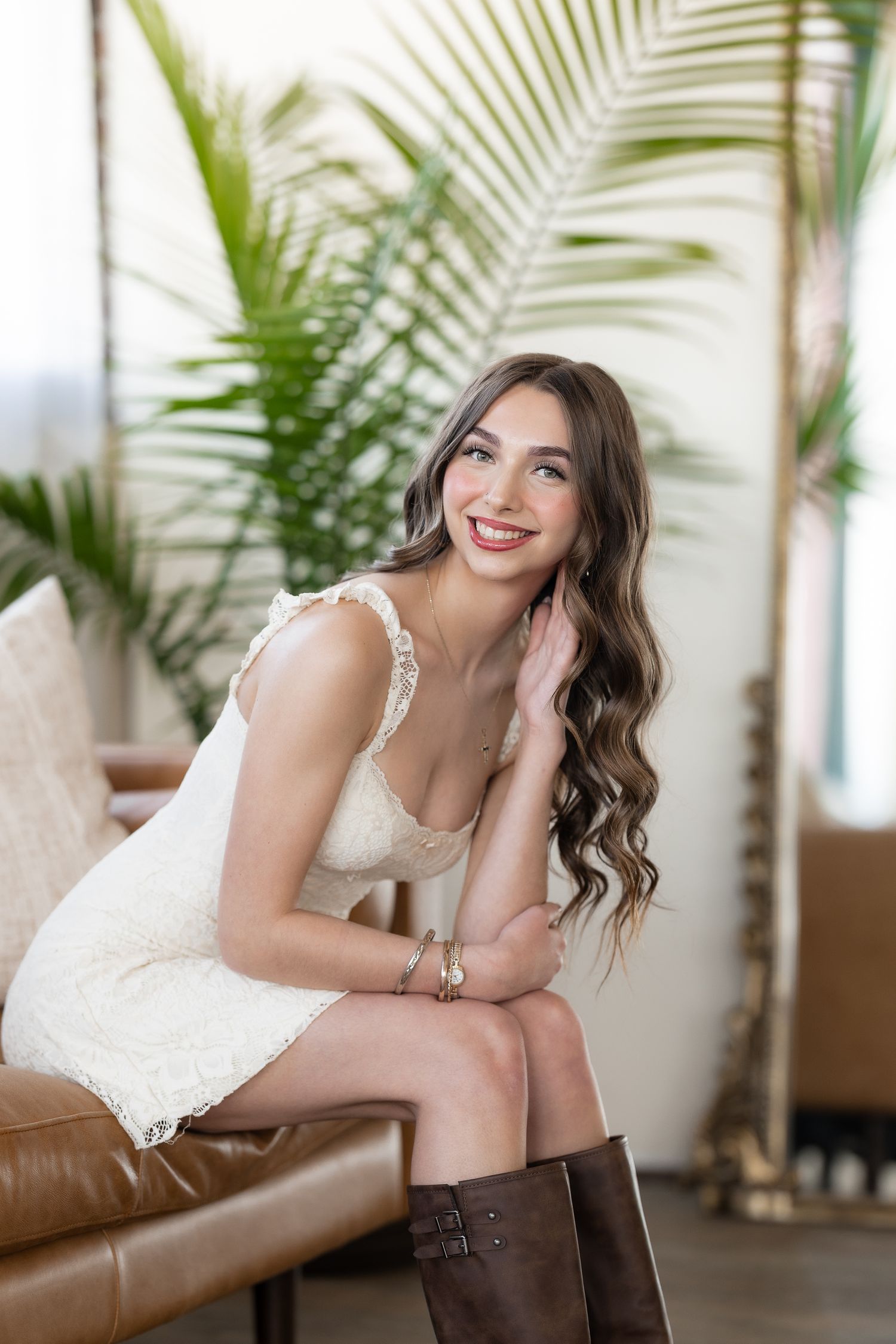 Young woman in white lace dress and brown boots sitting on leather sofa, smiling, with tropical palm plant background.