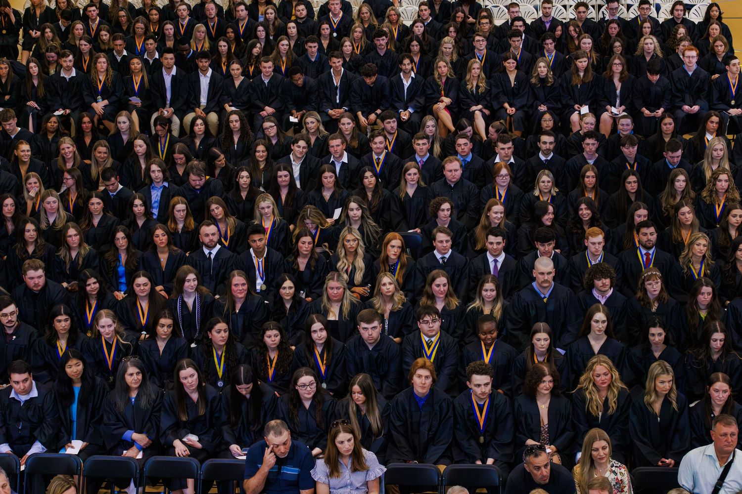 Large group of students in dark graduation gowns seated in rows during a formal graduation ceremony.