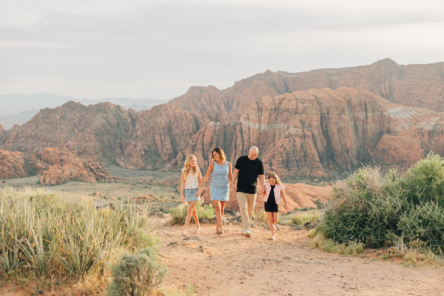 Family of four walking through a stunning red rock desert landscape with dramatic mountain formations in the background.