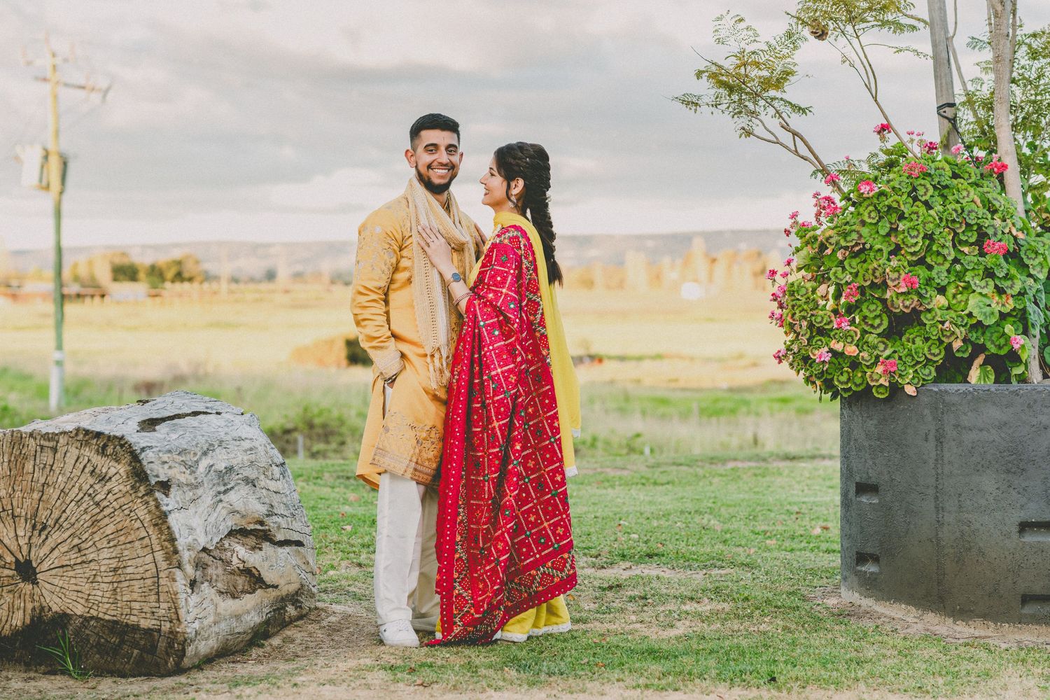 South Asian couple in traditional attire posing outdoors, man in gold sherwani and woman in red lehenga, scenic field backdrop.