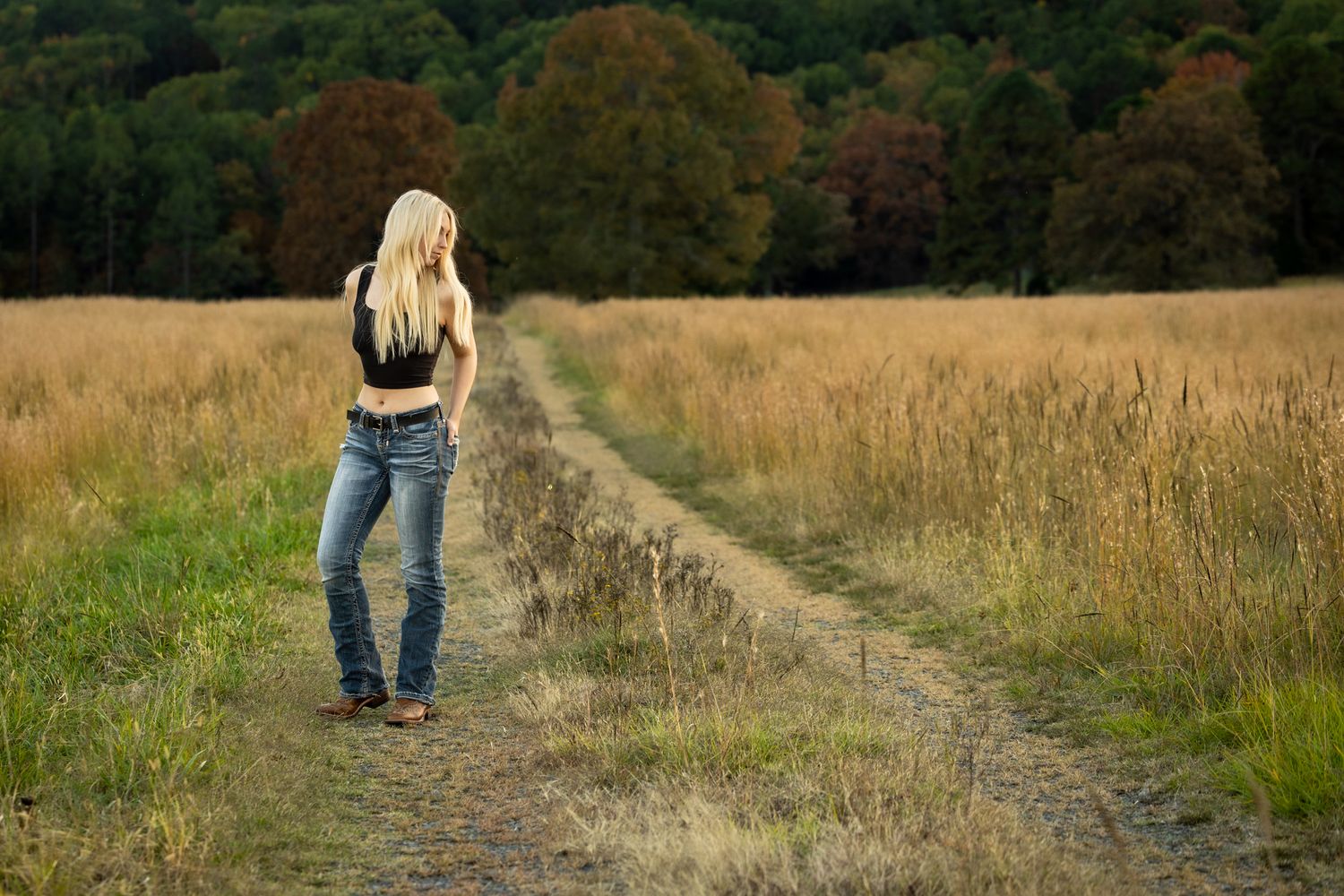 Blonde woman in jeans and black top standing on a rural dirt path surrounded by golden fields and autumn trees.
