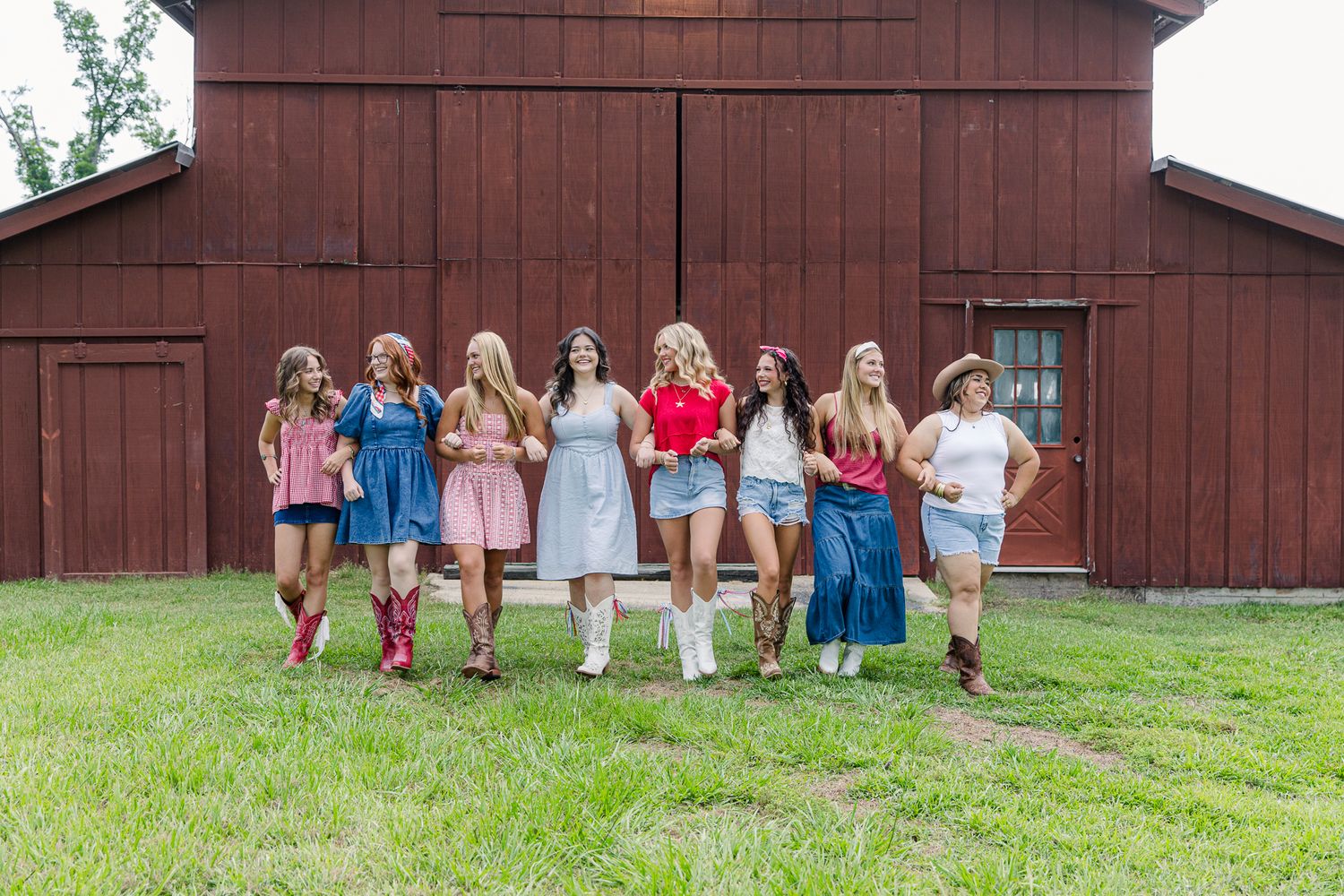 Eight young women in colorful country outfits pose together in front of a rustic red barn on green grass.