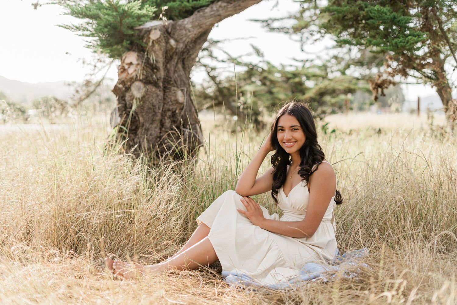 High school senior in a cream dress sitting beneath a tree at Laguna Lake Park during a San Luis Obispo senior photo session.