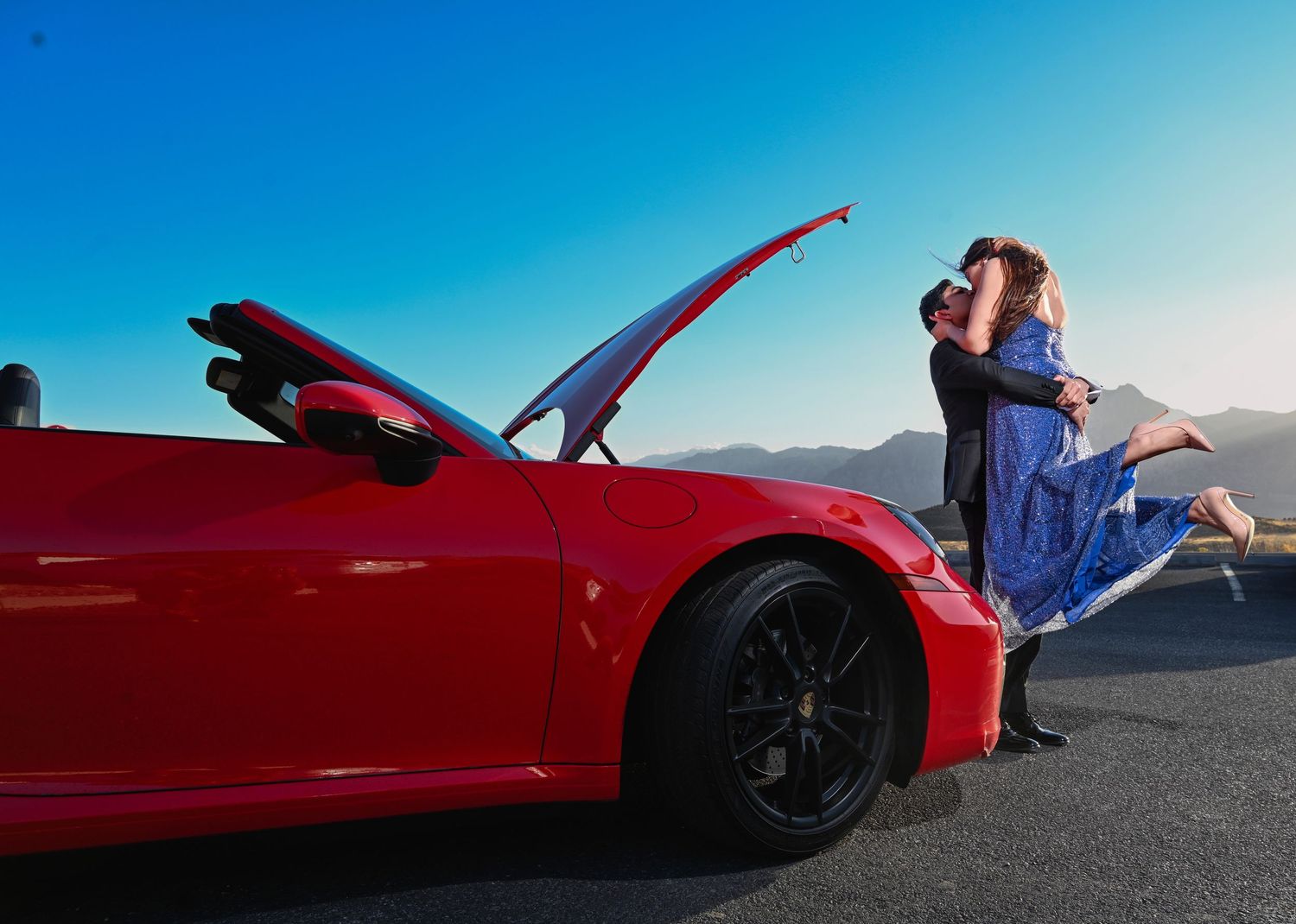 Couple embracing romantically beside a red sports car with open hood against a clear blue sky.