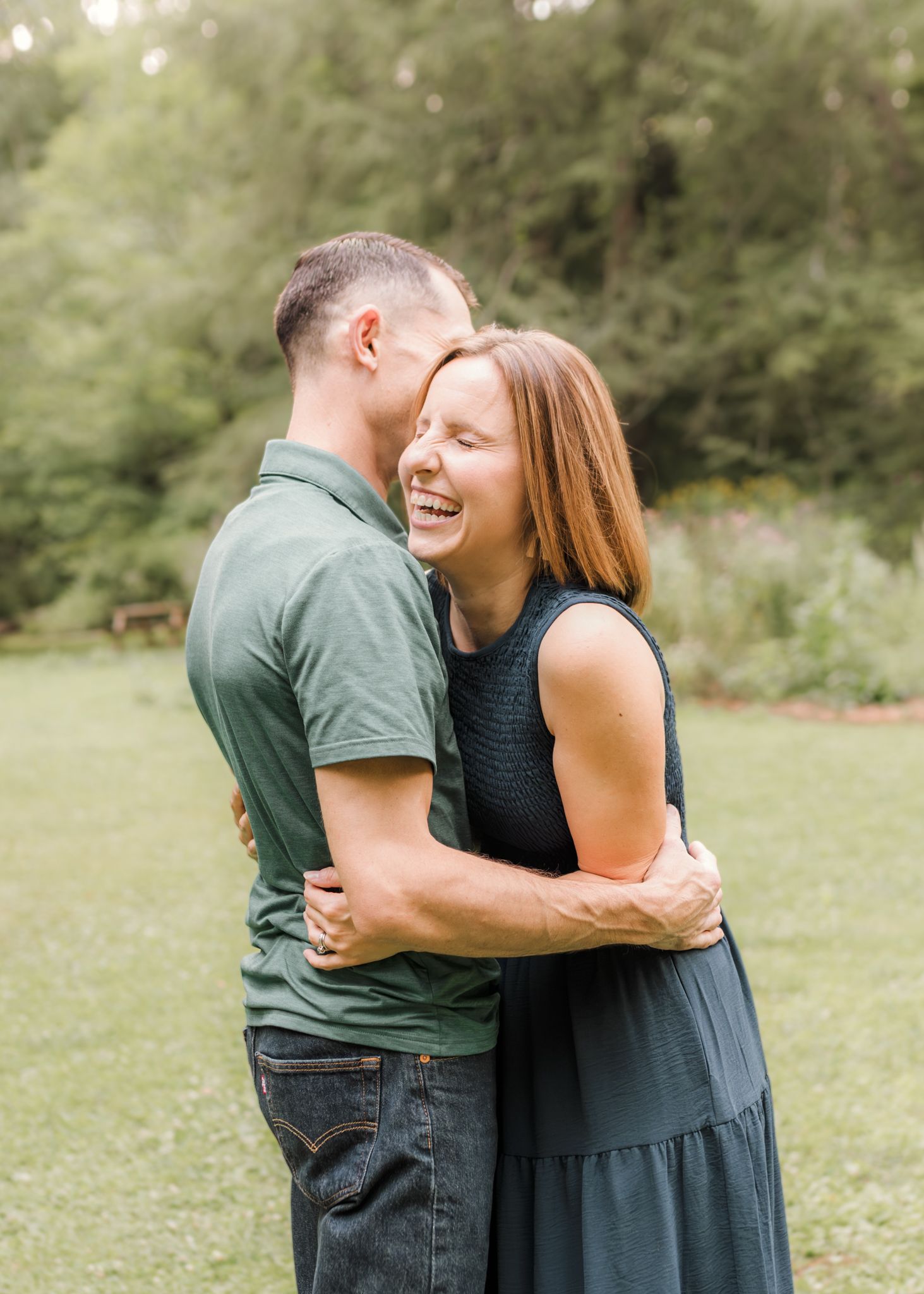 A joyful couple embraces and laughs together outdoors in a lush green park during a romantic engagement photo session.