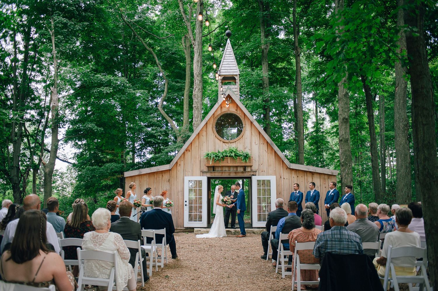 Outdoor wedding ceremony at a rustic wooden chapel surrounded by tall trees with guests seated in white chairs.