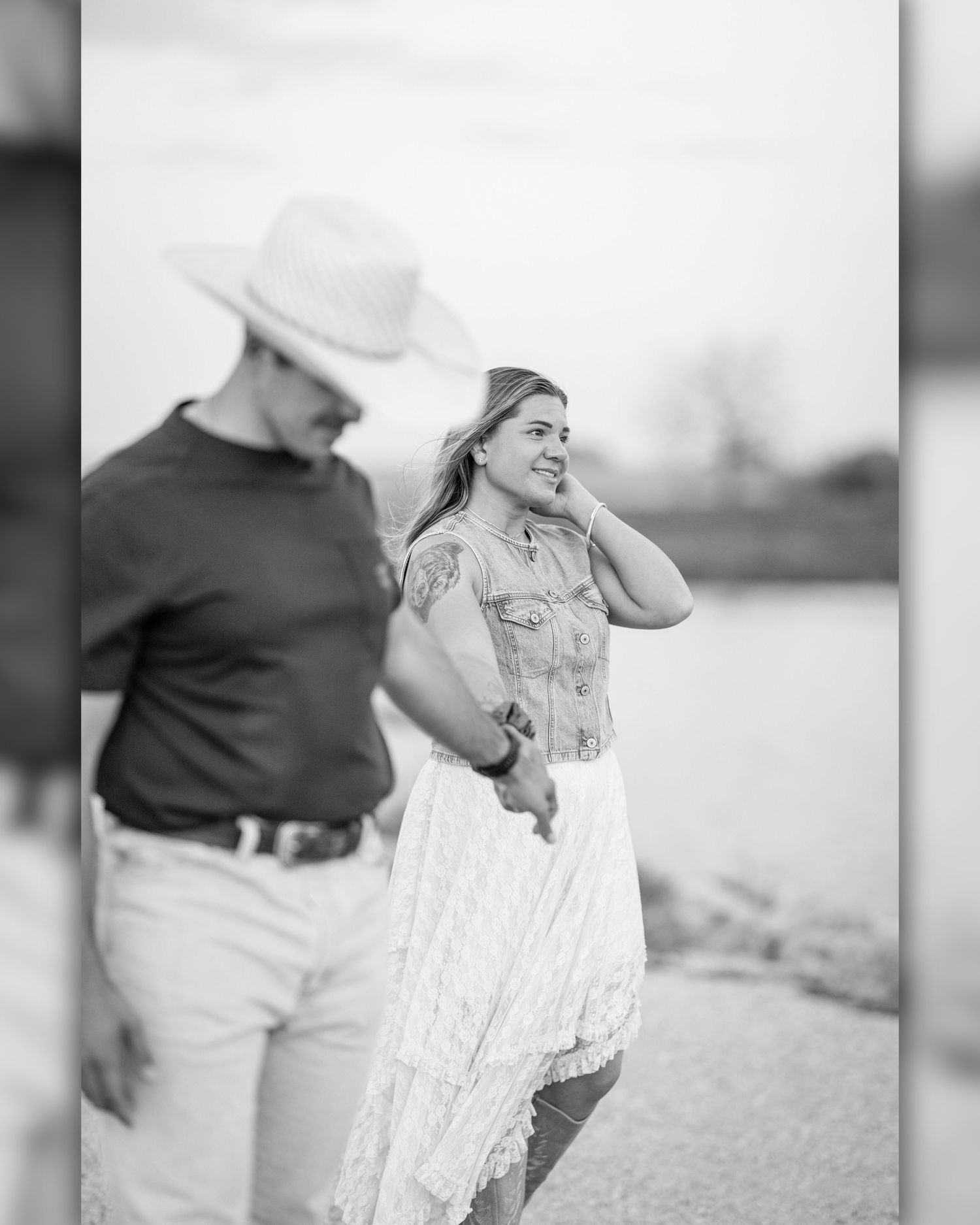 Black and white photo of a couple holding hands outdoors near water, woman in white dress smiling, man in cowboy hat.