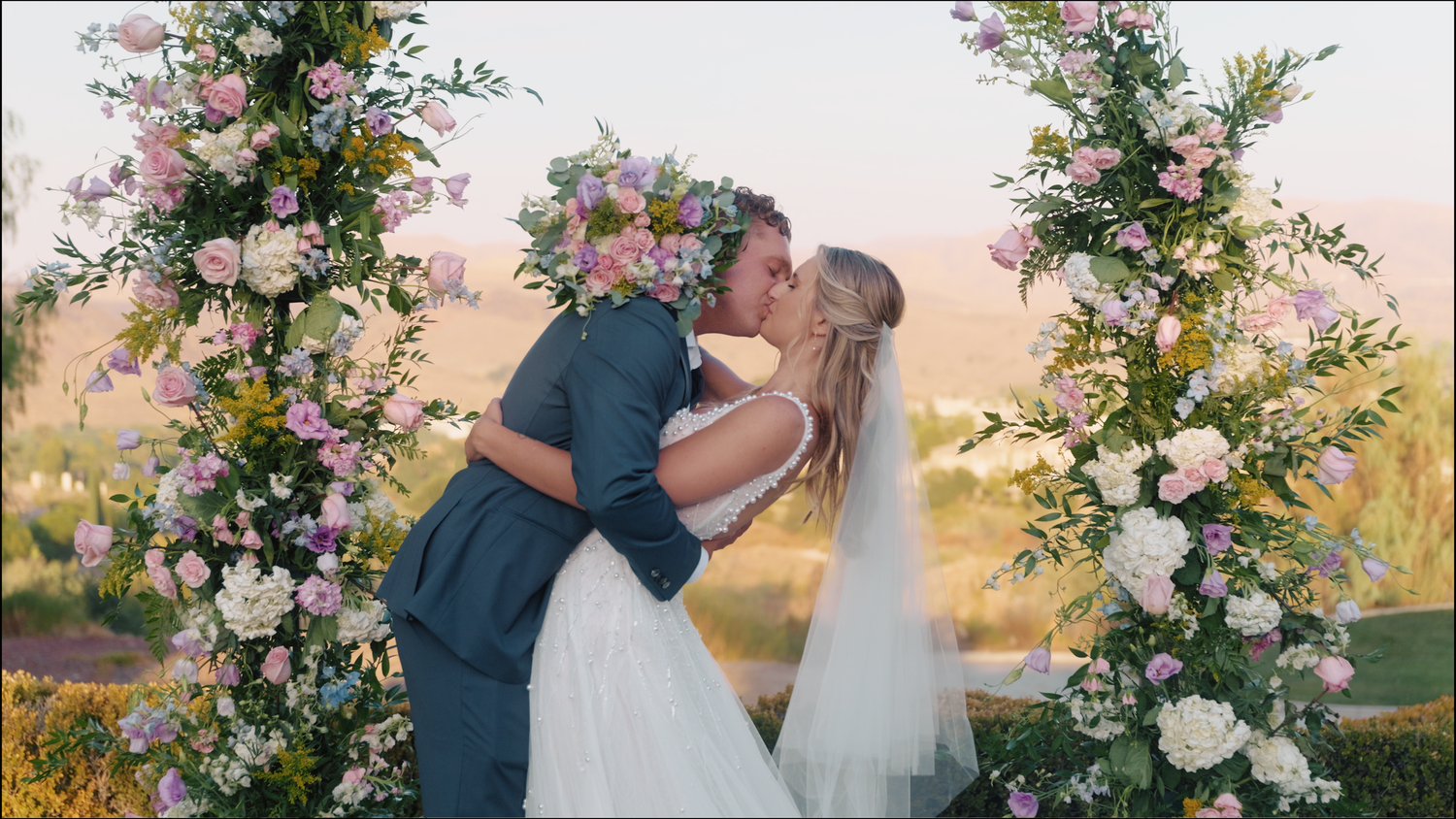 A bride and groom share a romantic kiss under a floral arch adorned with pink and white flowers at an outdoor wedding.