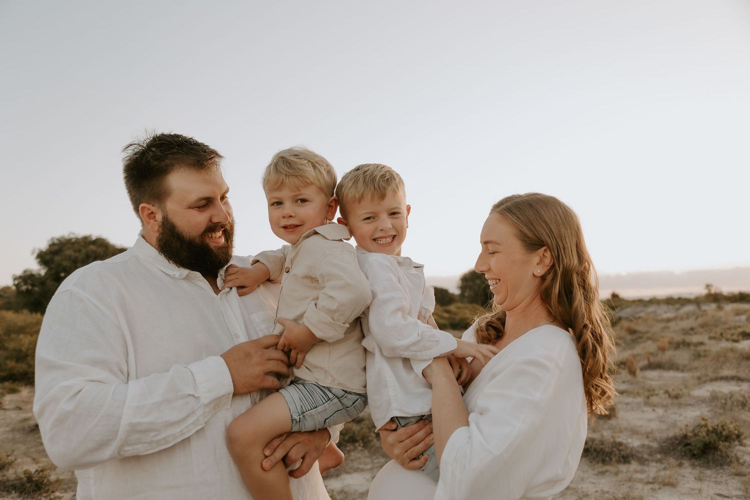 Happy family of four in white outfits, parents holding two young boys, smiling outdoors in a sunny sandy setting.