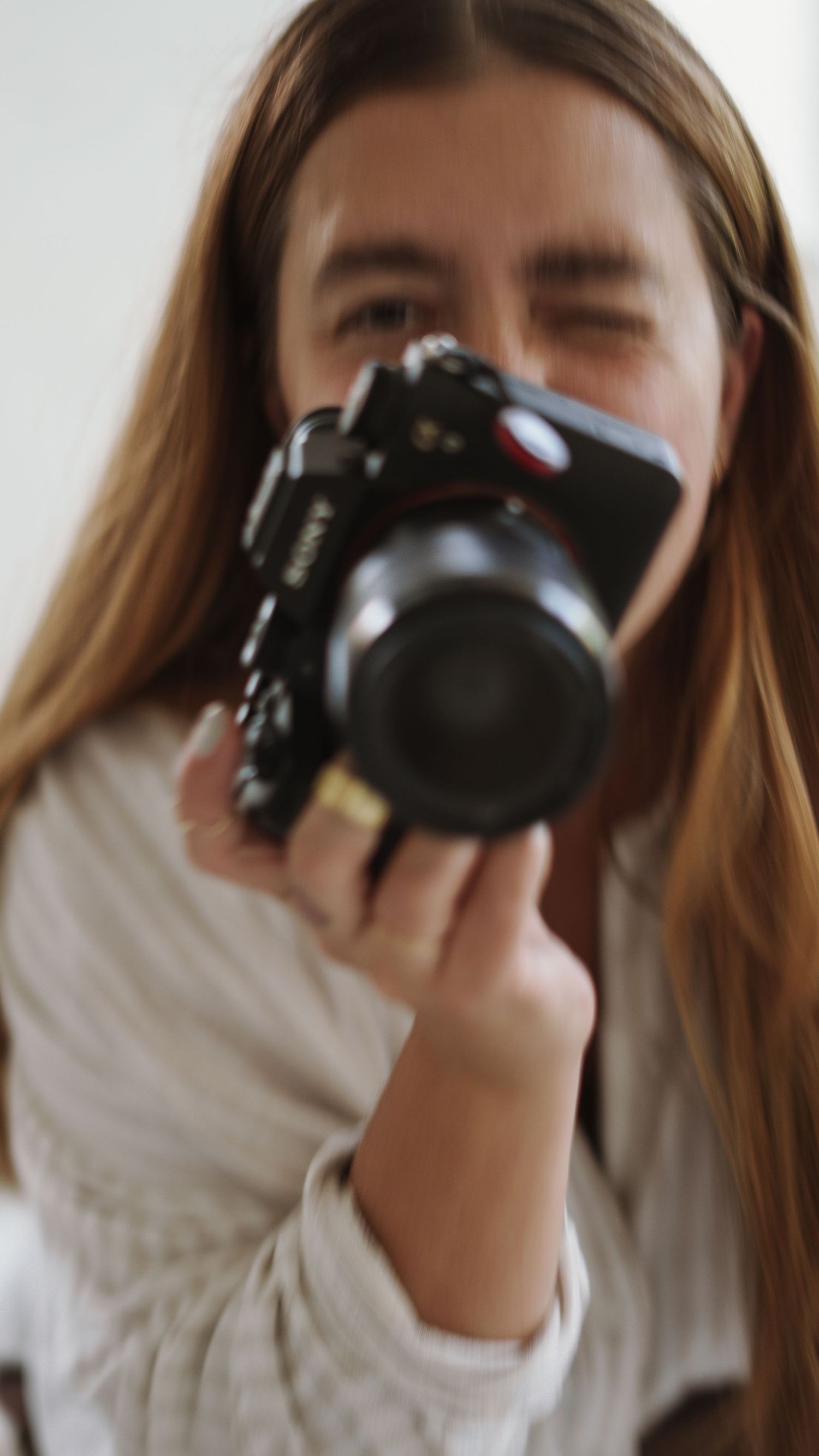 Young woman with long brown hair holding a black DSLR camera up to her eye, photographing indoors.