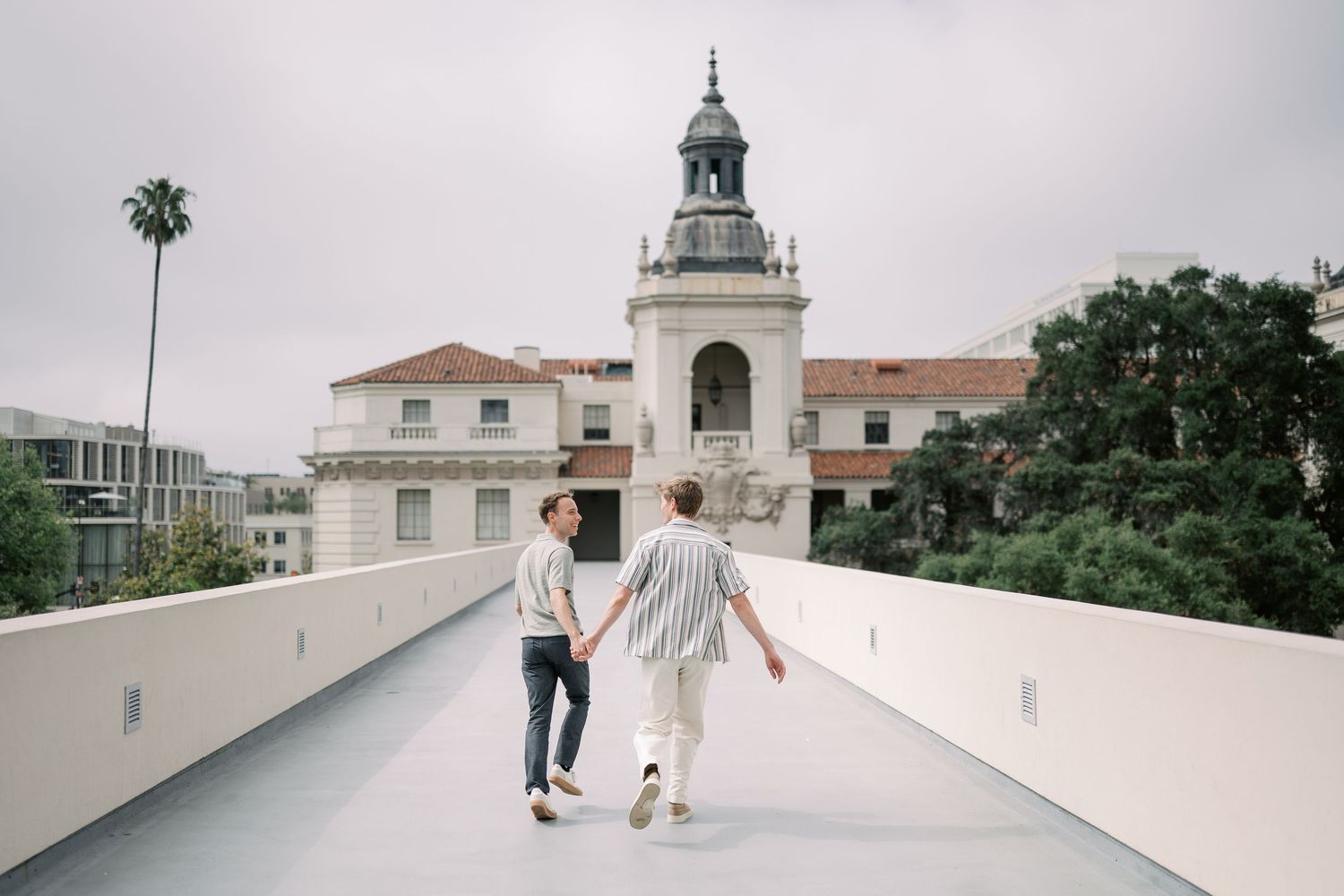 Couple holding hands walking toward historic Pasadena City Hall building with palm trees and cloudy sky.