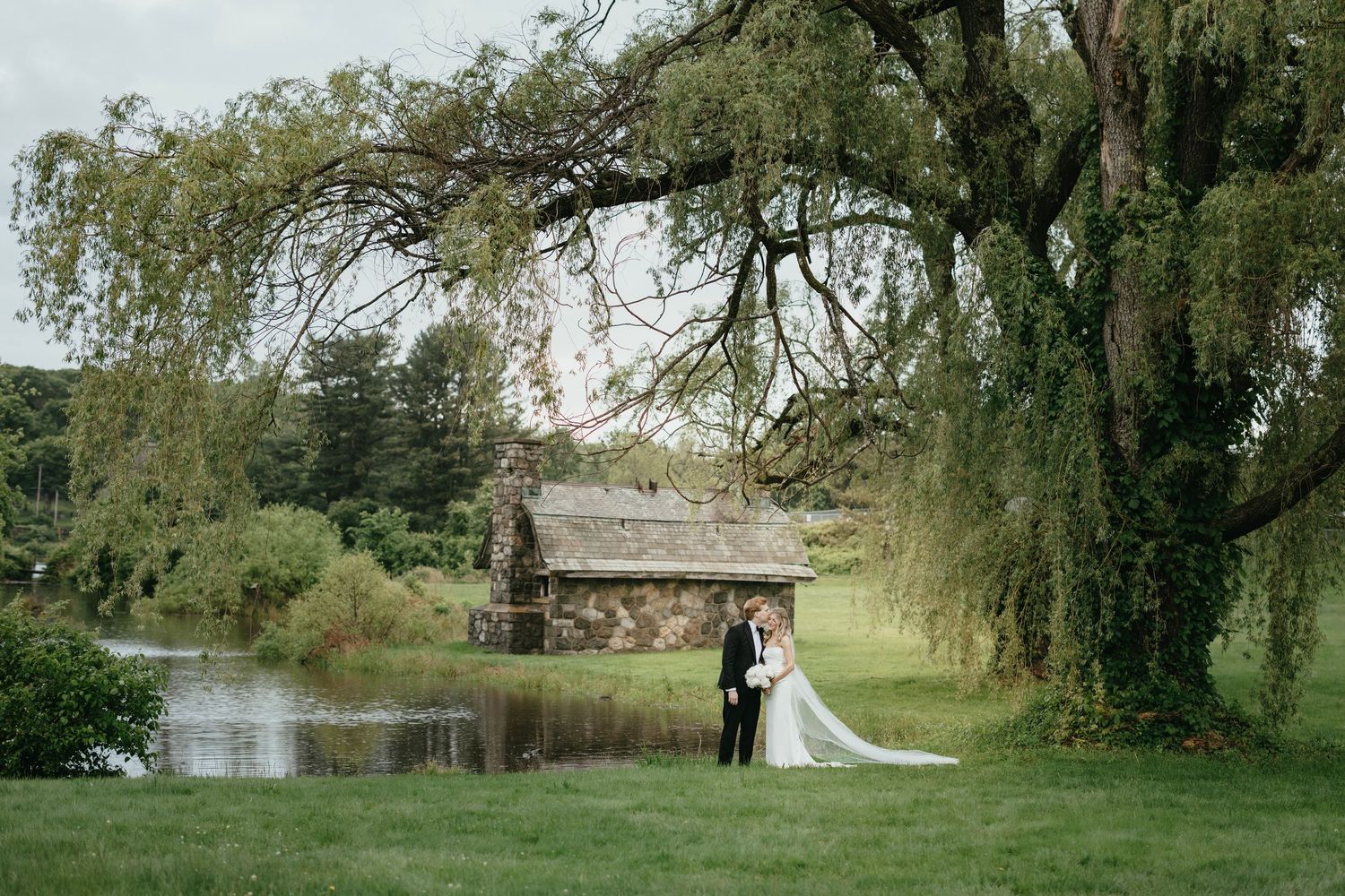 Outdoor wedding ceremony takes place on lawn in front of grand stone castle with Gothic architecture under cloudy skies.
