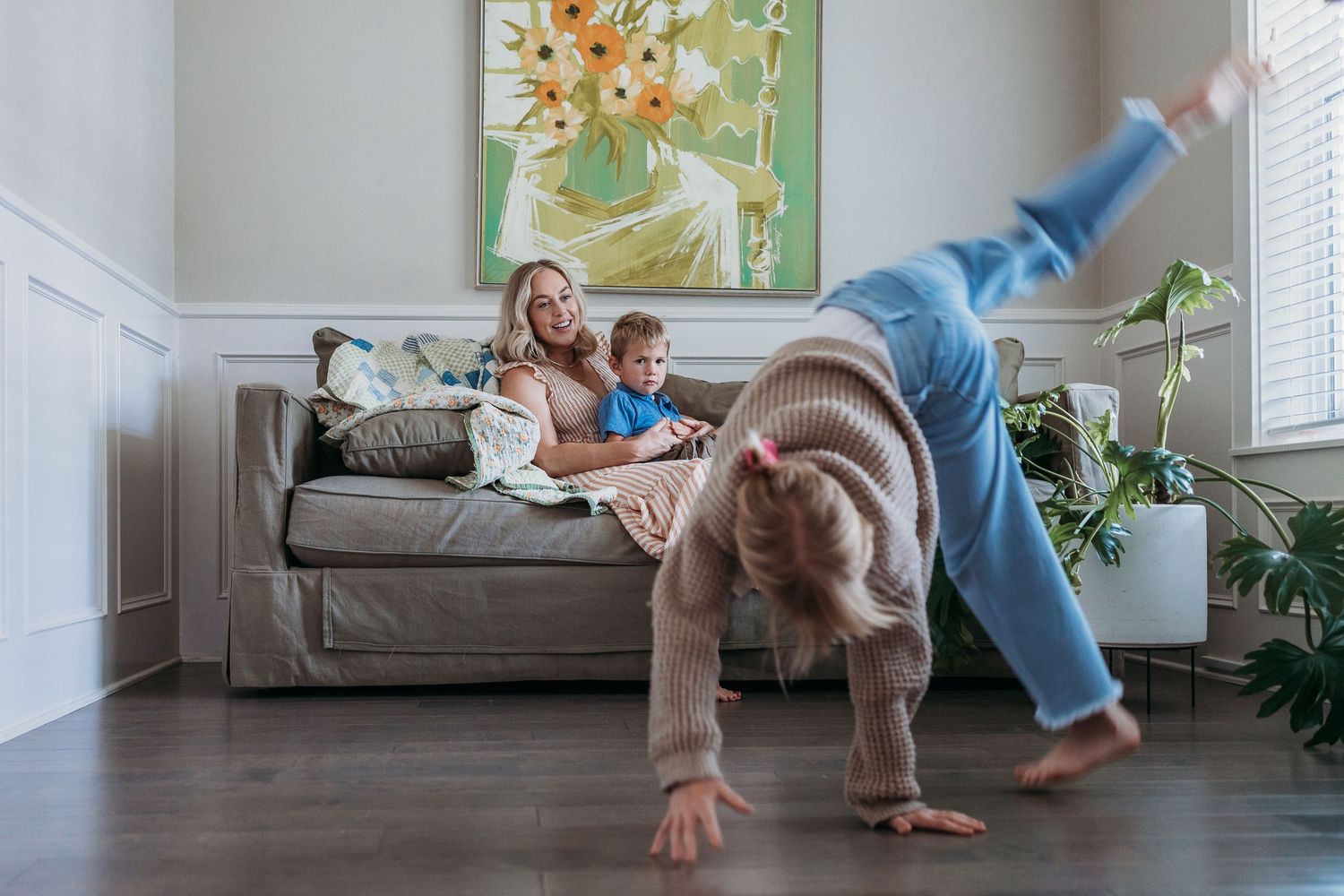 A person performs a cartwheel sequence in a bright living room with houseplants and a gray couch.