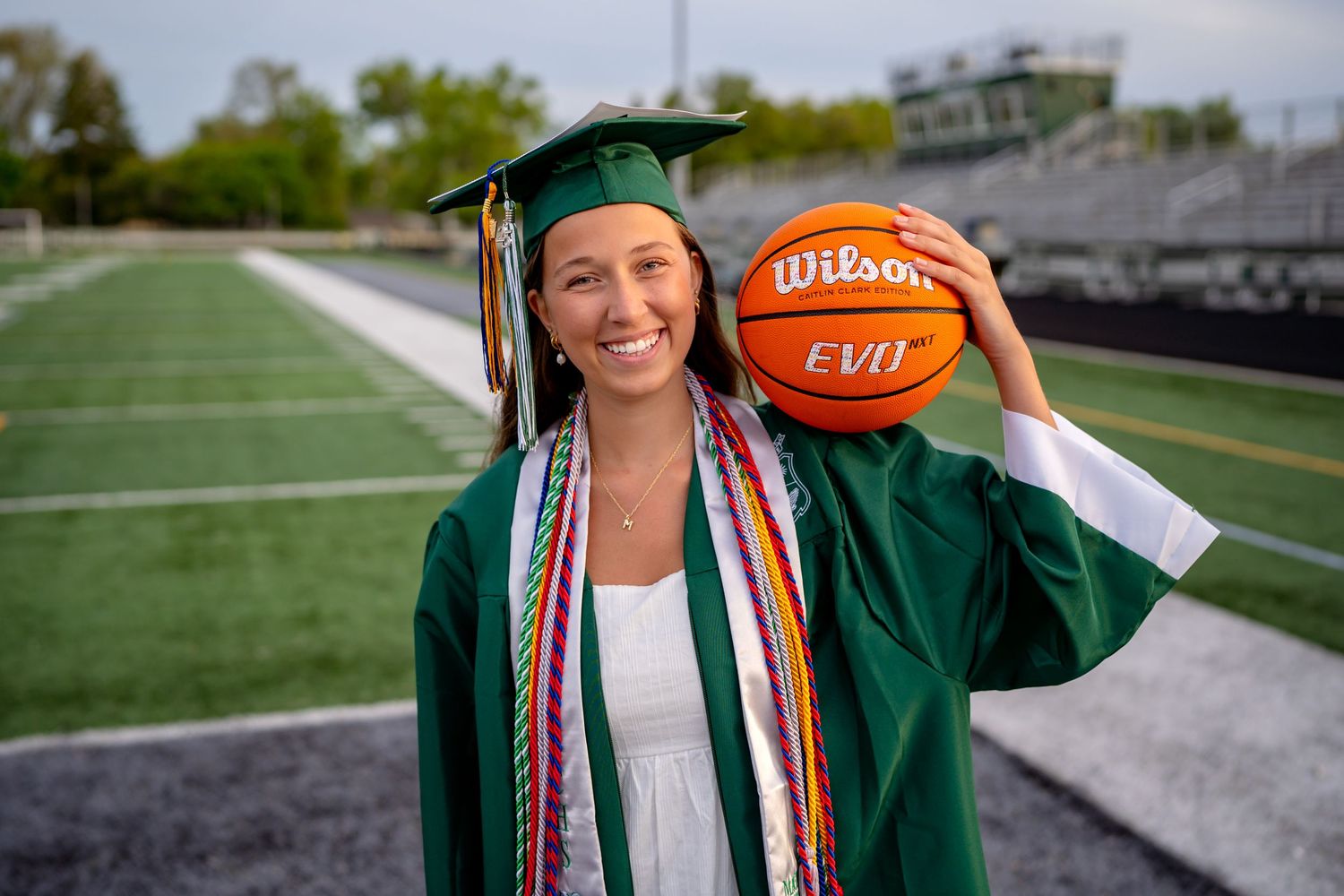 Graduate in green cap and gown holds a football while posing on an athletic field during graduation photos.
